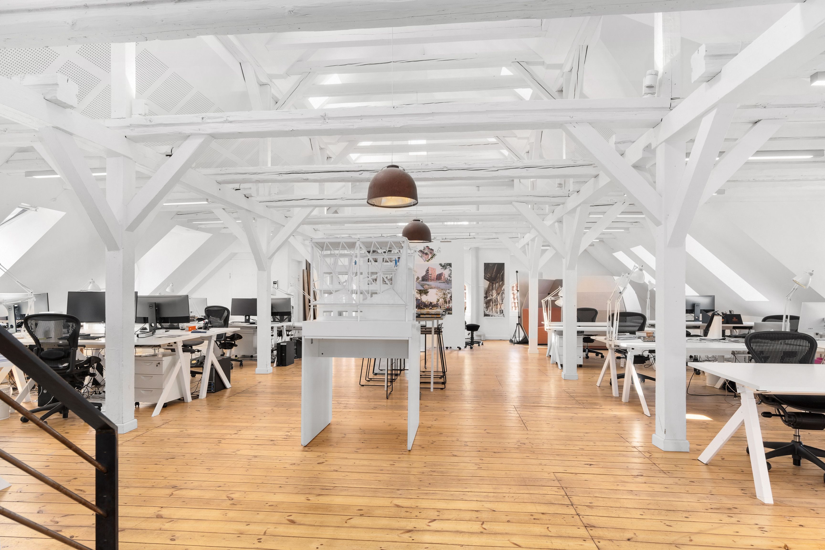 Kontorlokaler med højloft, lyse loftsbjælker og moderne møbler, hvide skriveborde og stole, og en stor loftsvinduesglas.