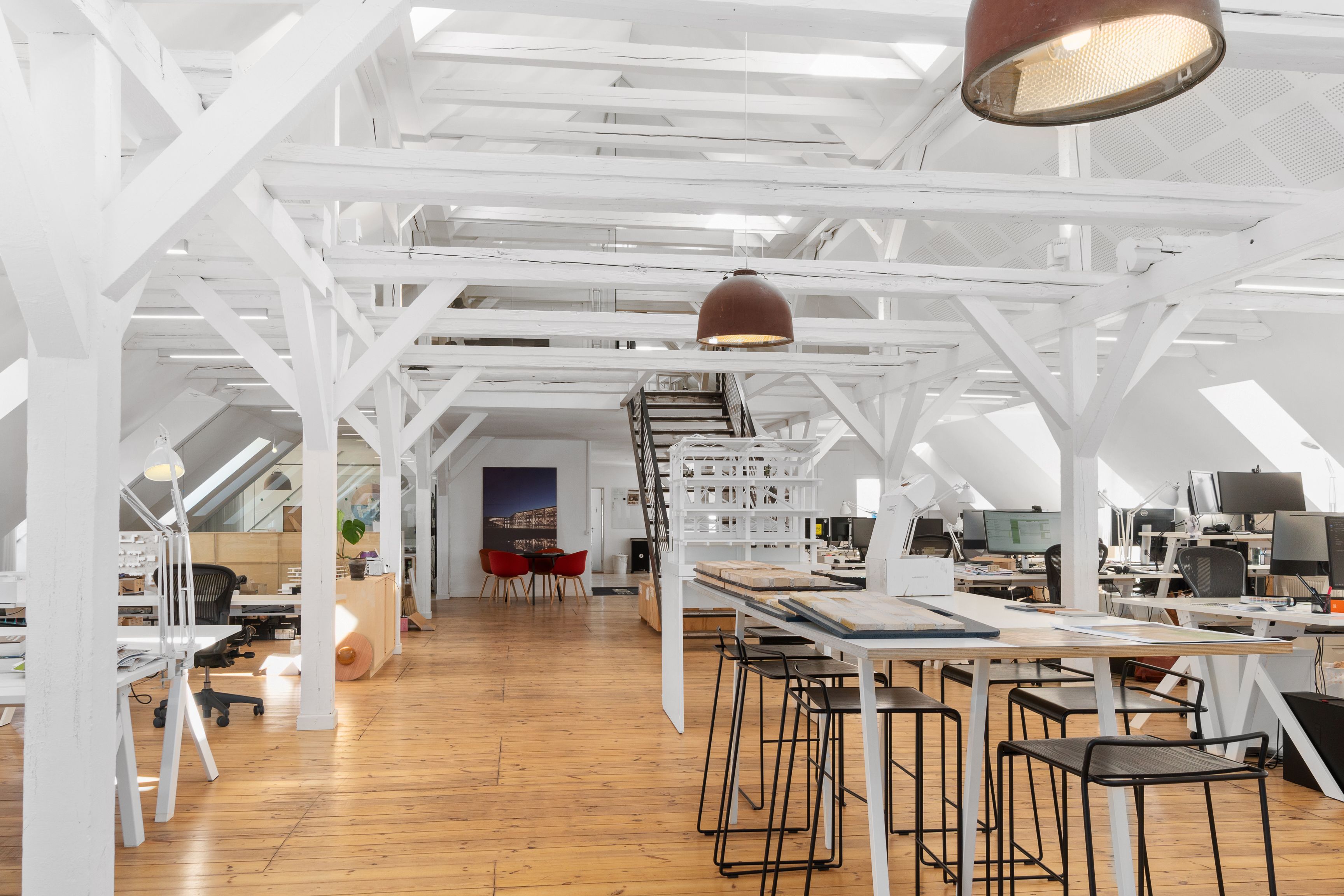 Kontor med højloft, lyse bøjle og lyse trægulve, hvor skriveborde og stole er placeret, og der er en stige med hvide trin