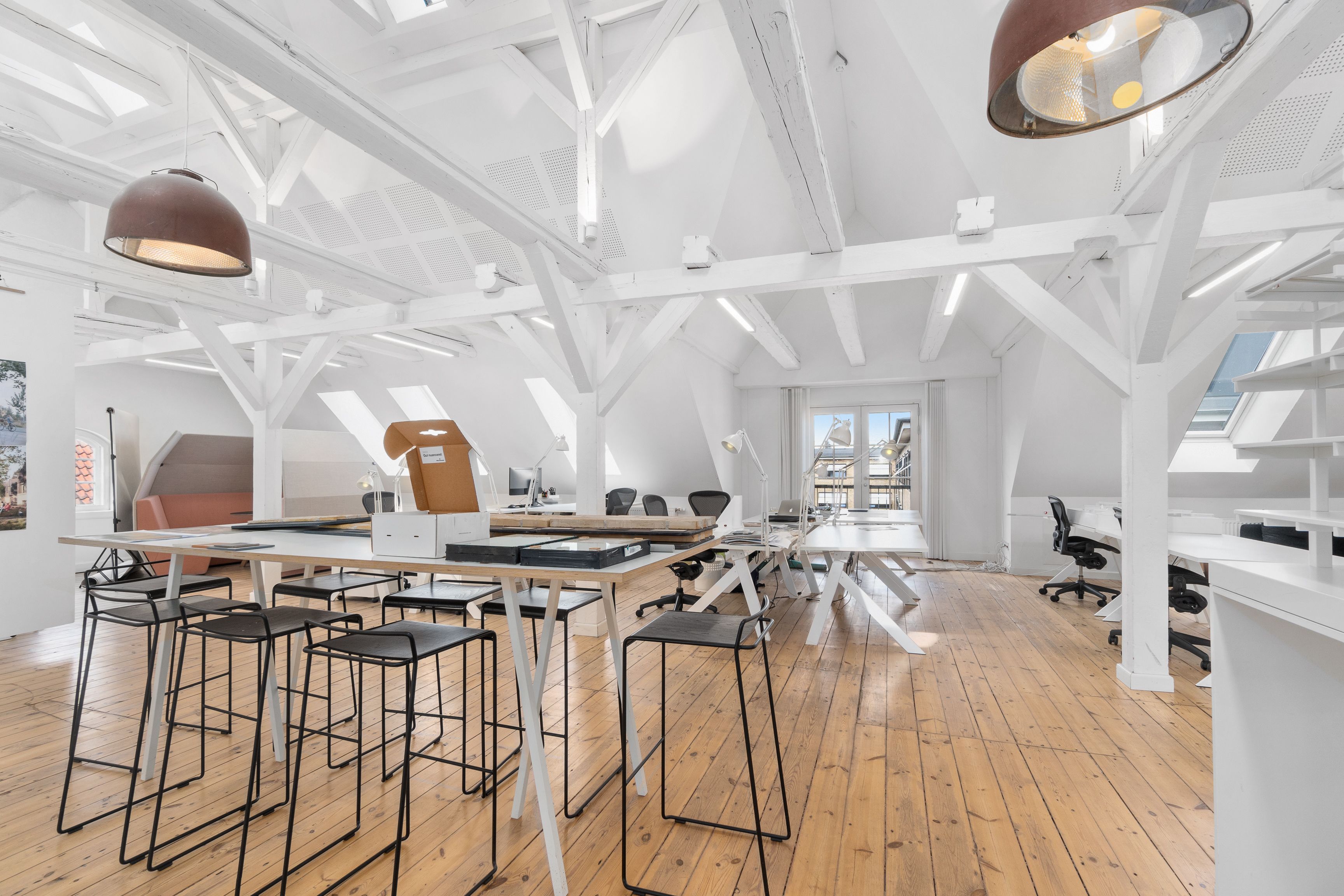 Kontor med højt loft og store vinduer. Moderne bord og stole. Bjælker og loftsvinduer. Slanke og høje lysekroner. Trægulv og minimalistisk indretning.