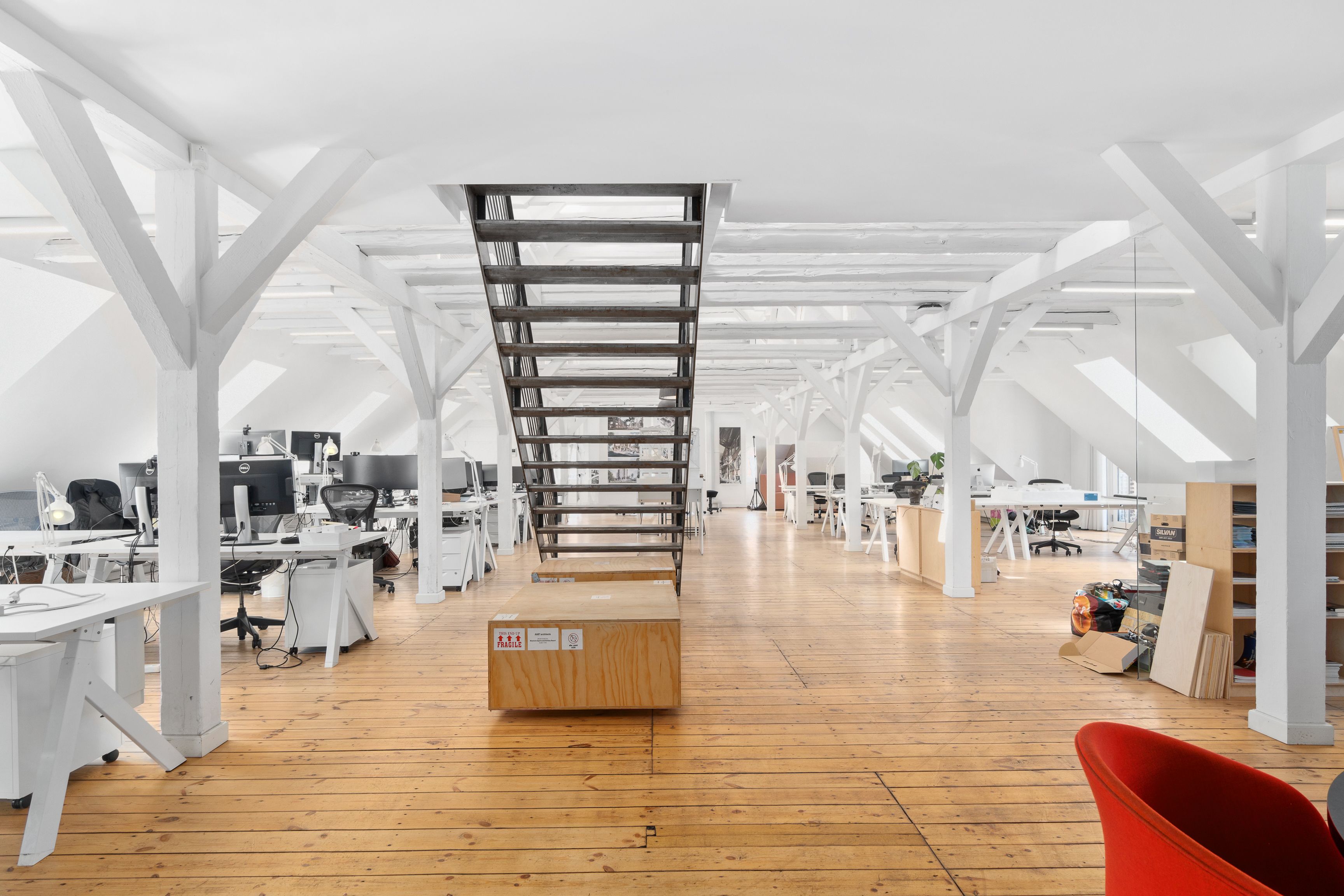 Kontorområde med lyse lofter, hvide bjælker, lysegul gulv og moderne møbler inkl. skriveborde, stolpe og et rød stol.