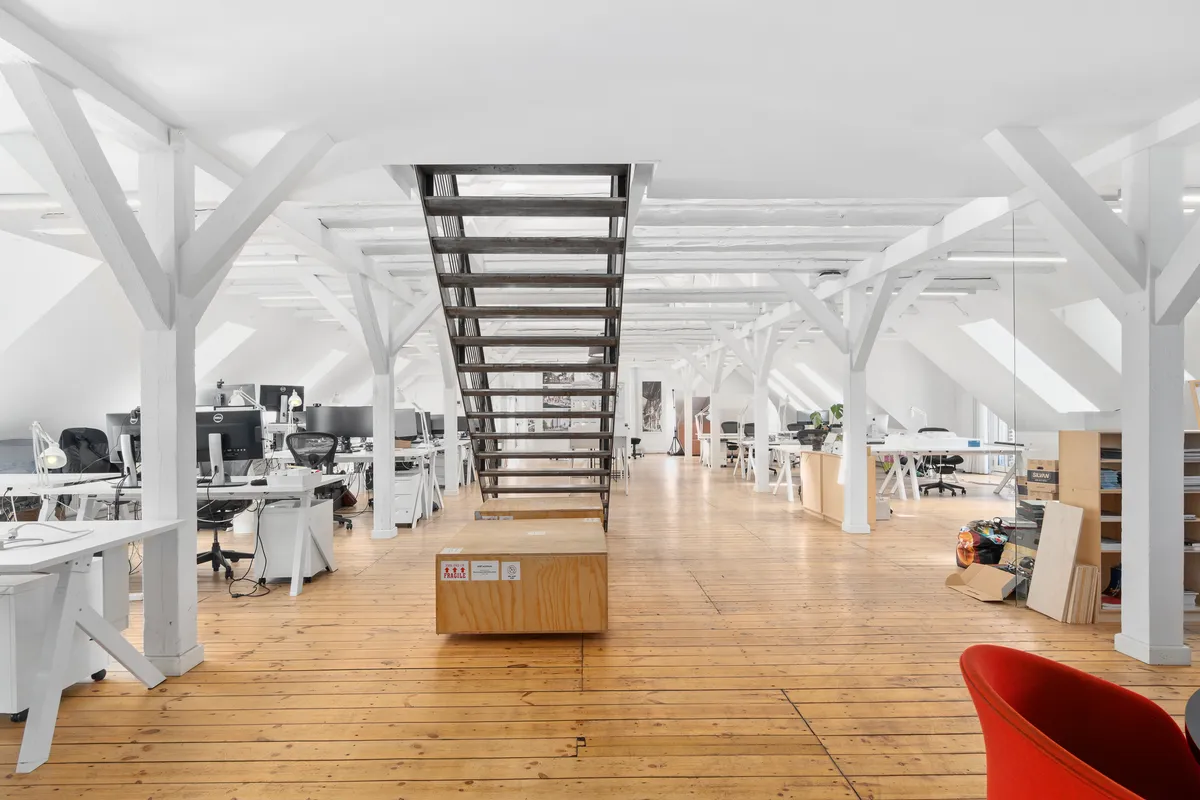 Kontorområde med lyse lofter, hvide bjælker, lysegul gulv og moderne møbler inkl. skriveborde, stolpe og et rød stol.