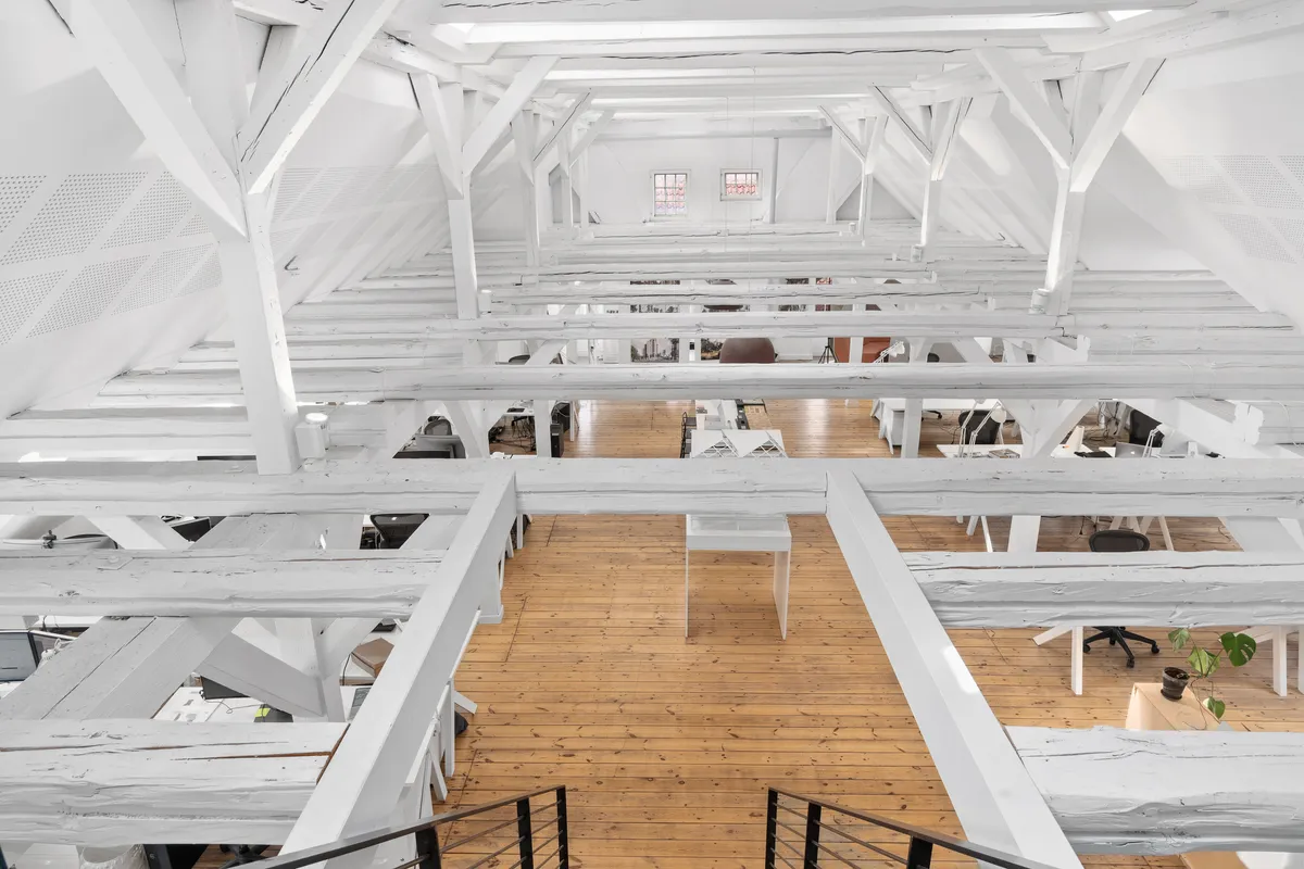 Kontorlokaler med hvide bjælker og trægulv, flere bordplader, stole og computere i rummet, der er indrettet med moderne møbler og faciliteter