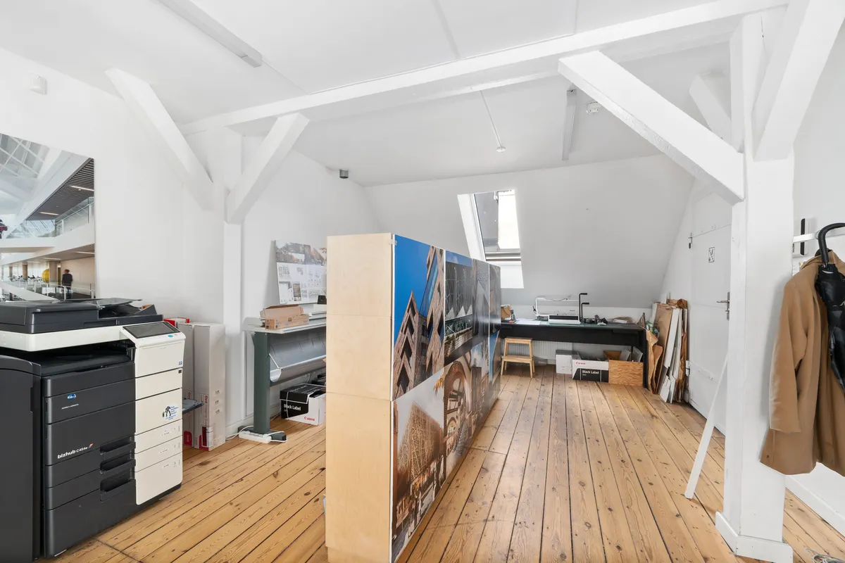 Kontor med lyse lofter og bjælker, printere, papkasser, stole og skrivebord med computer og skærm