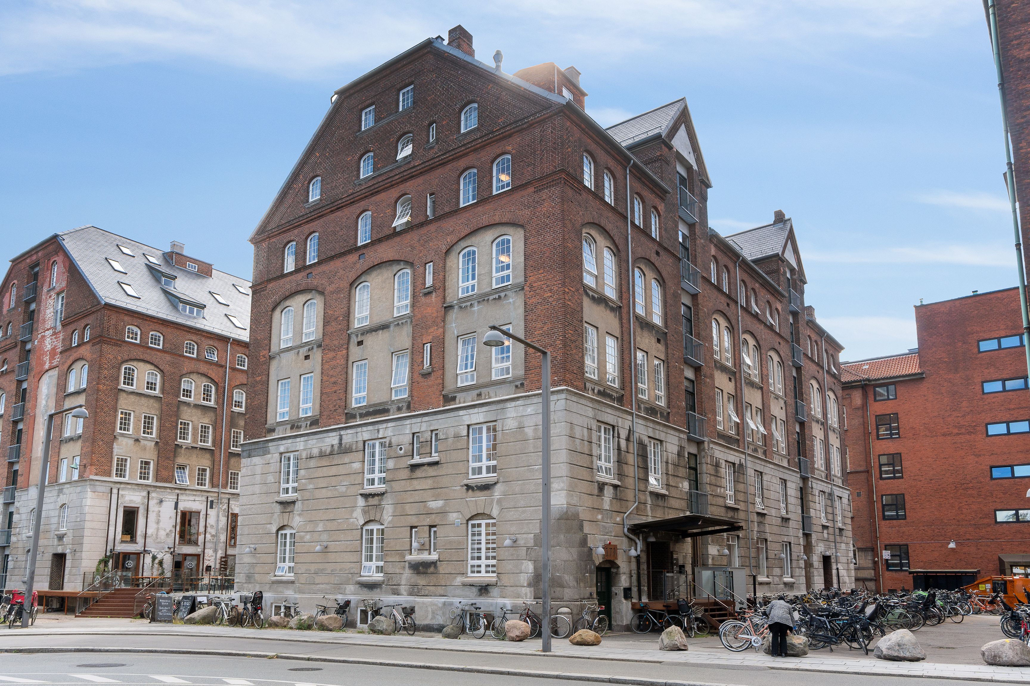 Lejlighedsbygning med røde mursten og flere vinduer i København. Foran bygningen er der parkerede cykler og en person står ved en cykel. Der er også stier med bænke og sten langs gaden.