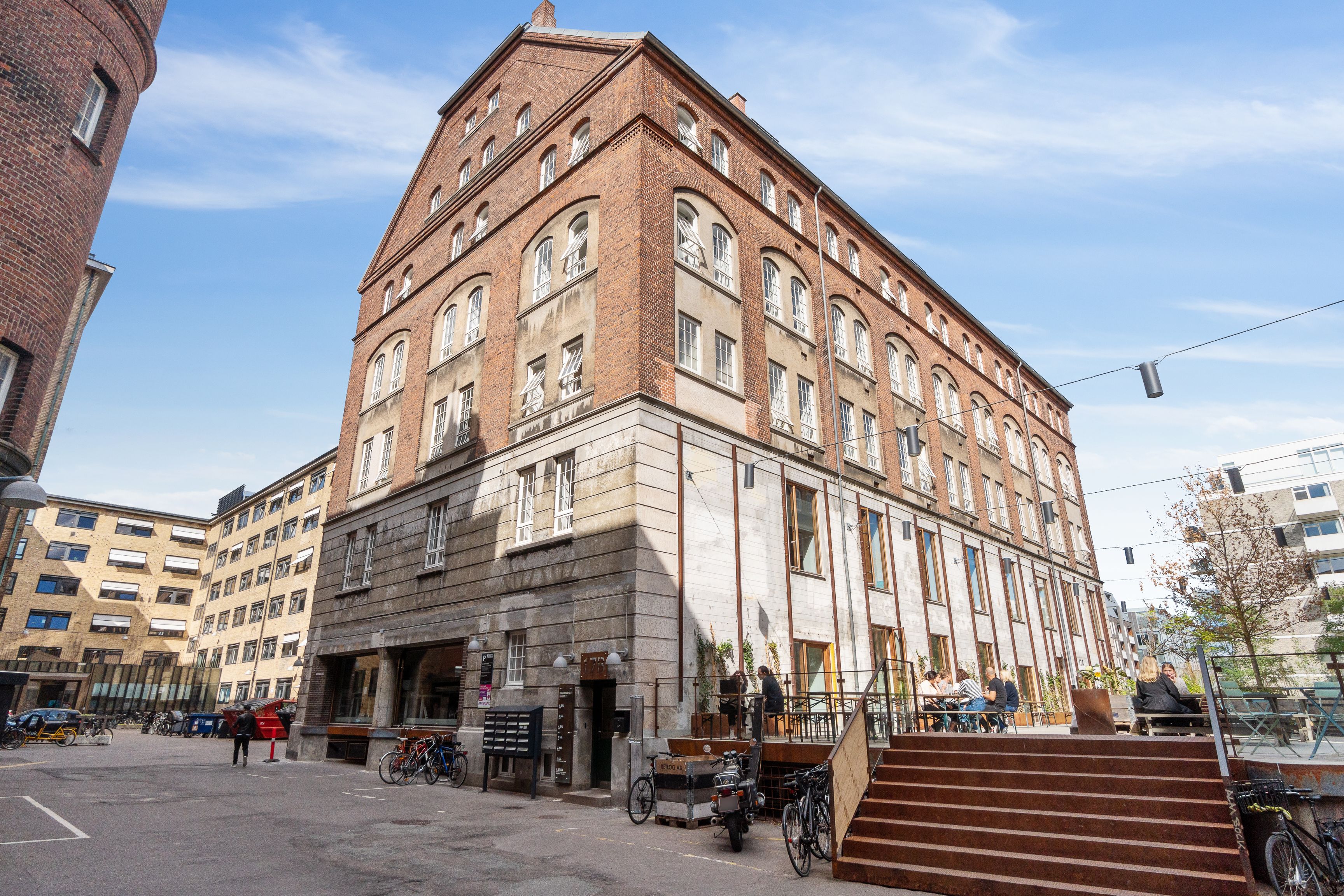 Moderne lejemål i rødmalet bygning med terrasse, flere cykler og stedet for caféer og restauranter med gæster i baggrunden