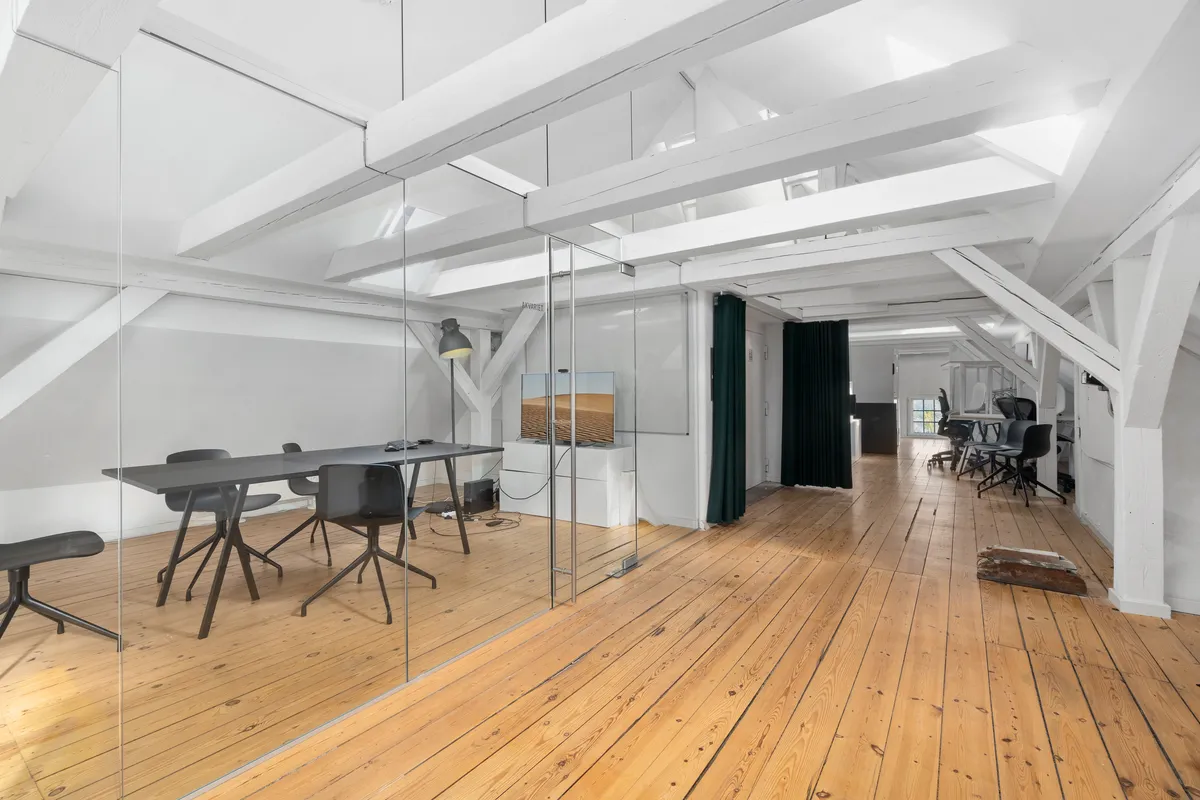 Kontor med højloft og lysegule gulvplanker, moderne møbler og en stor glasvæg med refleksioner af lamper og skriveborde