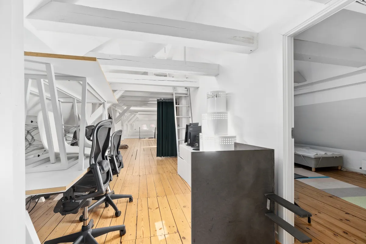 Kontorrum med højloft og bjælkeloft, møbler inkluderer skriveborde, kontorstolar og en hvid høj kabinet, samt en monitor og en stige i baggrunden