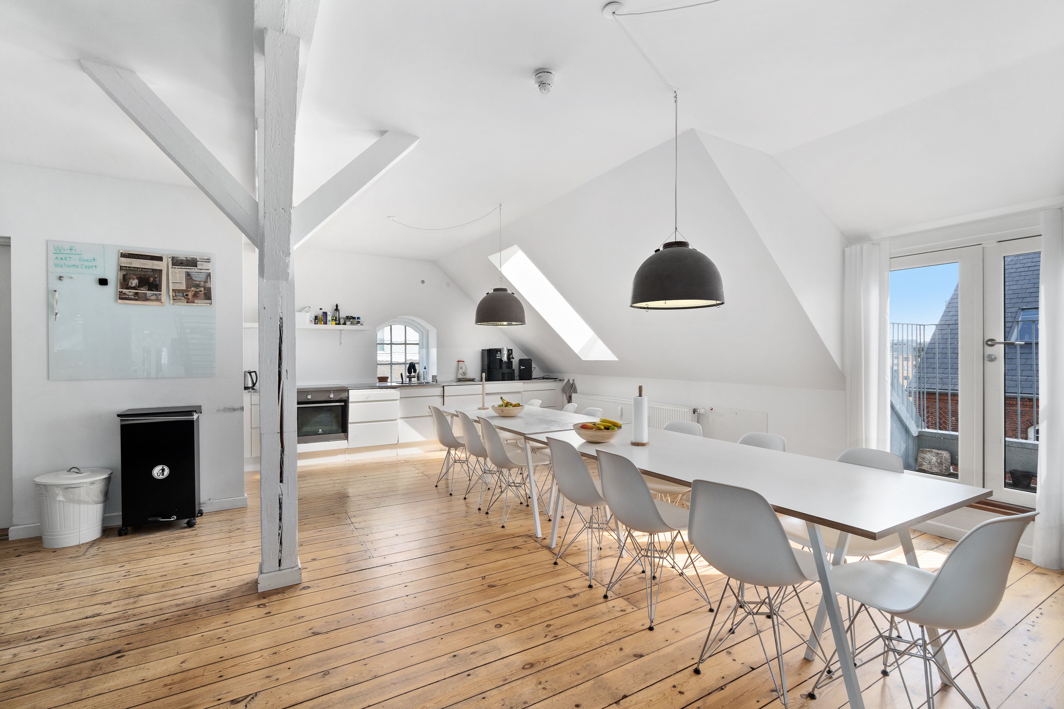 Kontorlokaler med højloft og lyse loftsvinduer og moderne trægulve. Langt hvidt spisebord med lyse stolene og pendullampe. Opbevaringsboks og høj, hvid ovn i køkkenets hjørne.
