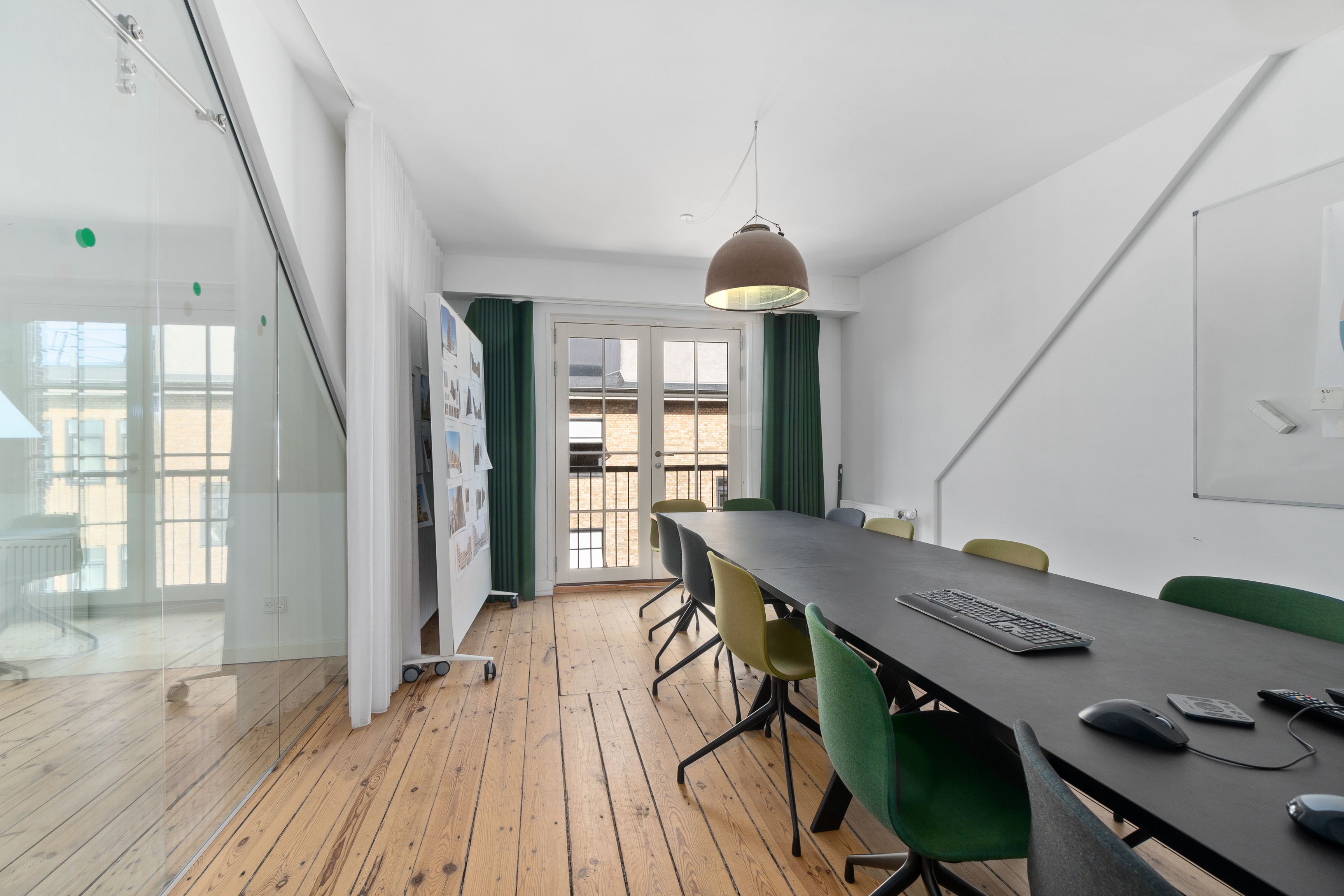 Moderne mødelokale med massivt bord, grønne stole og skrivebordsudstyr. Lampe hænger fra loftet og vinduer med grønne gardiner. Glasdøre med udsigt til balkon og bygning.