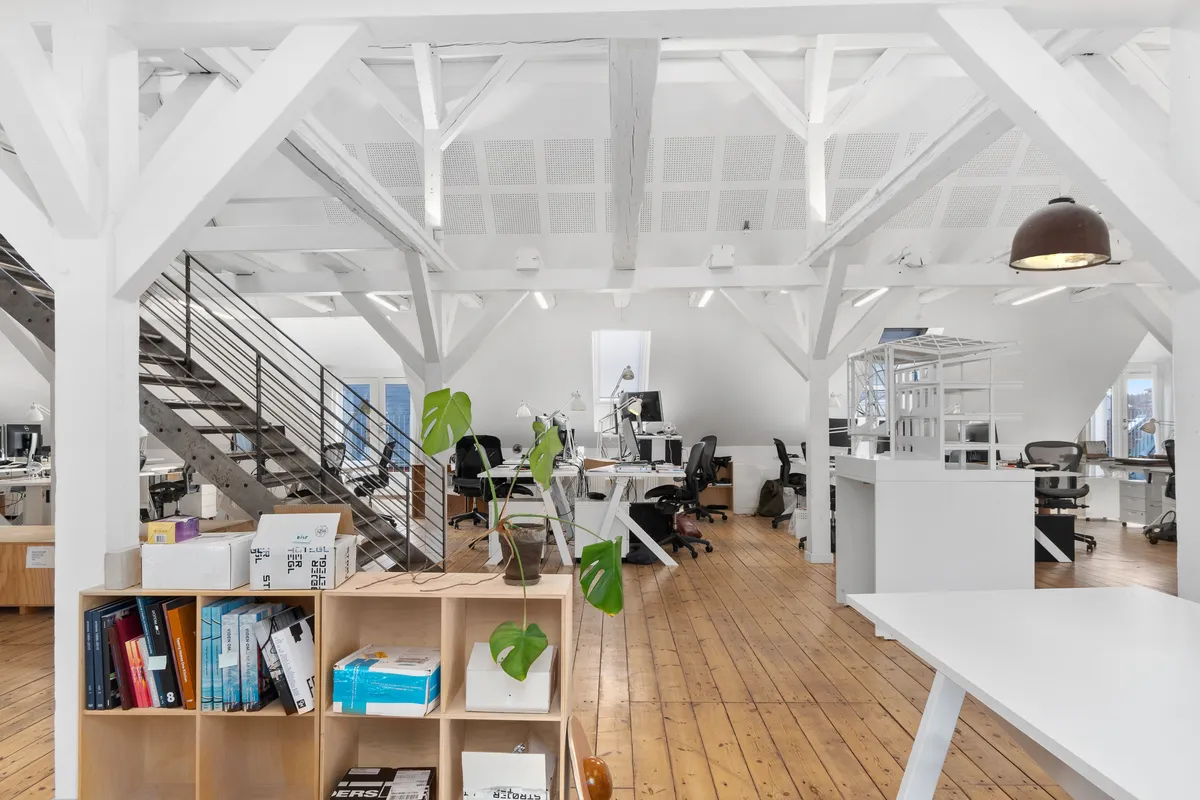 Kontorlokaler med højt til loftet, fritlagte bjælker, moderne skriveborde og plante. Værelse med høj gulvdekning og møbler og udstyr til arbejde.