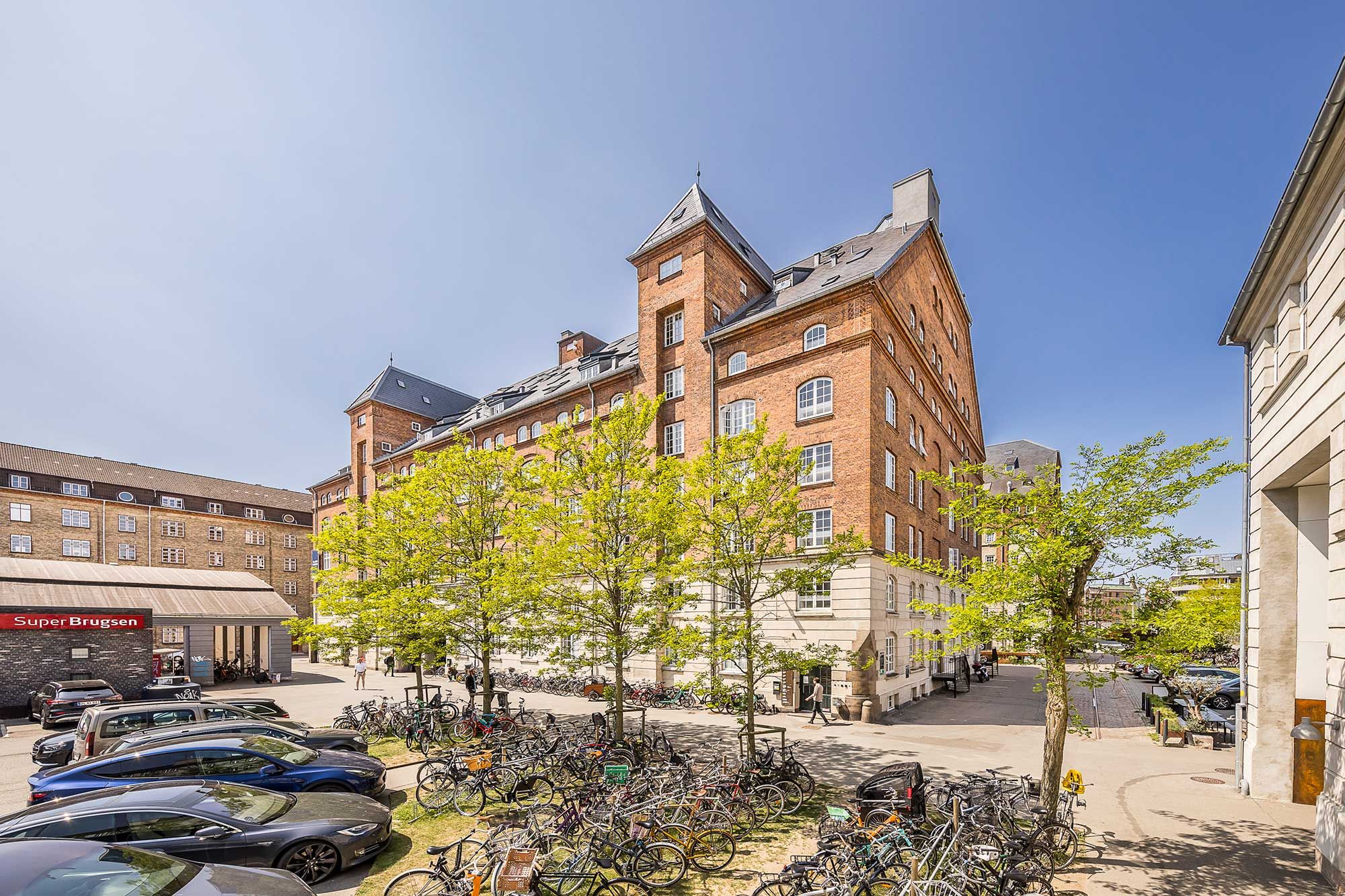 Parkerede biler og cykler foran en stor bygning med mange vinduer og bjælker