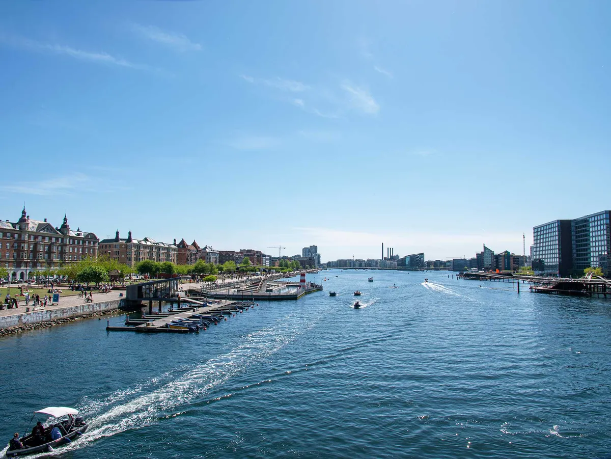 I baggrunden er der bygninger og en bygning med tårn. I midten er der et vandløb med både og små både. På vandet er der mennesker, og de er på en båd.