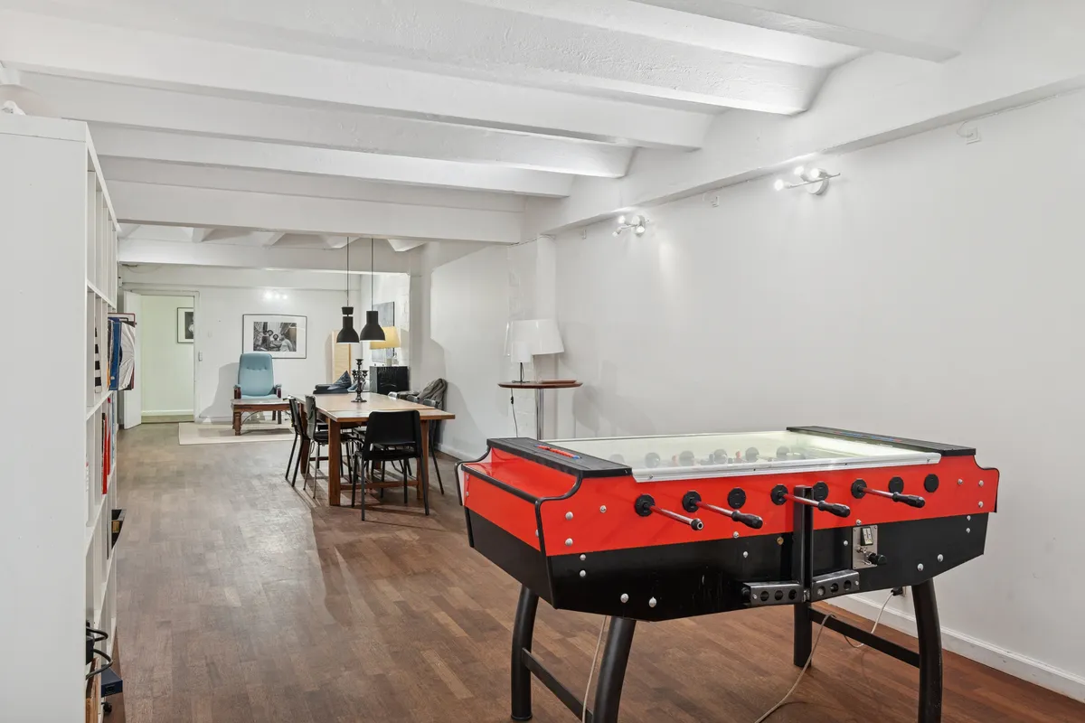 Hvidt og lysegult interiør med trægulv og loftsbjælker. Et stort bord med stole, en stol og en fodboldbord. En hvid lampe og et lys i loftet. En sofa og et bord med stol i baggrunden.