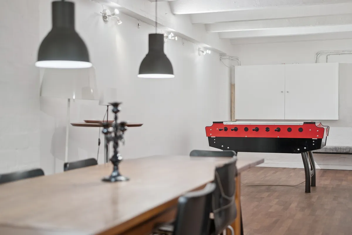 En kontorlokal med højt til loftet og lyse loftsbjælke. Væggene er hvide og lyse, og der er en gulvfodbejle og en rød og sort fodboldspil. Der er en lang træbord med to stole og en lampe i midten