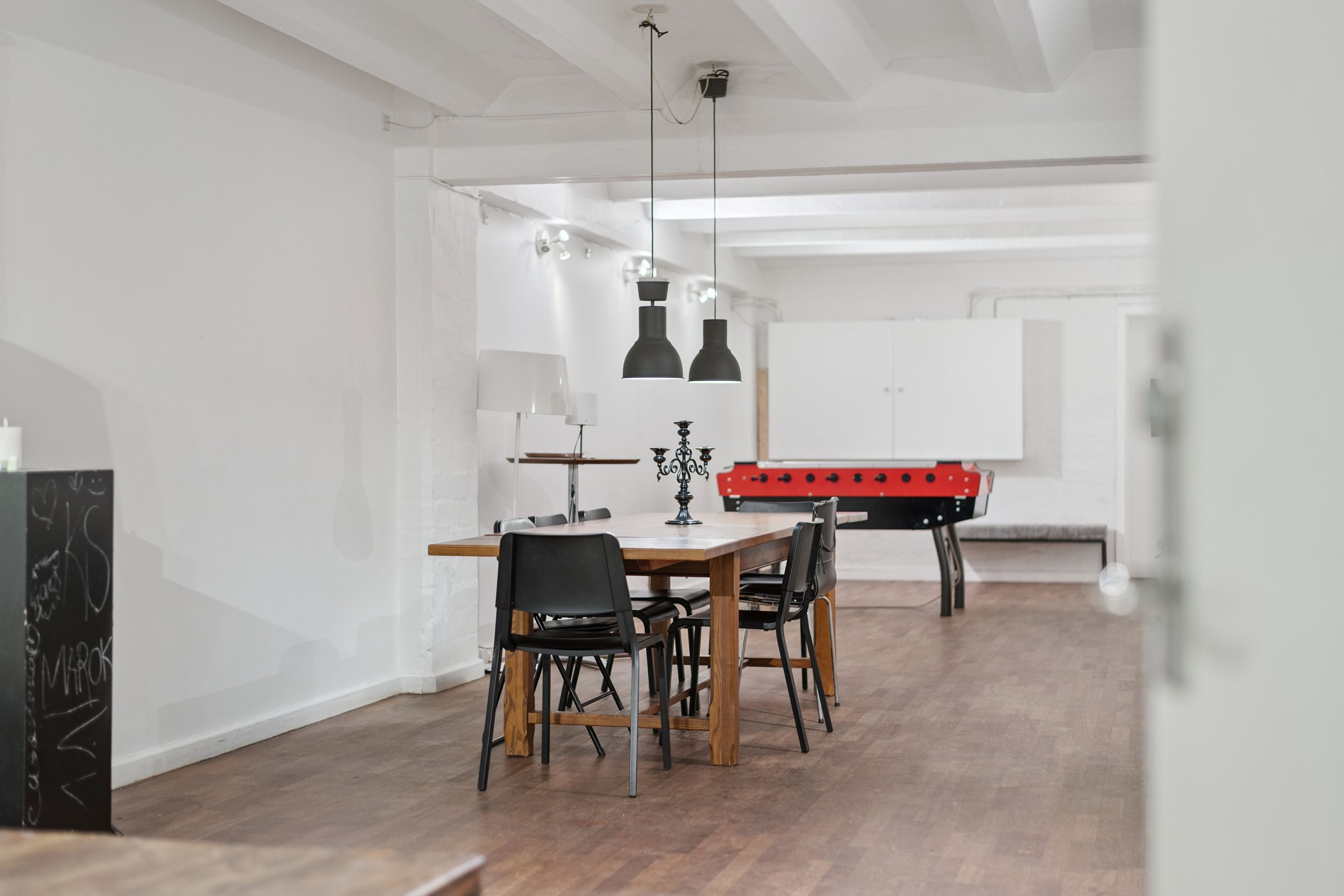 En stor stue med højt til loftet, hvor et langt bord og stolpelampe er i centrum, og en bordfodbold er i baggrunden, alt i et minimalistisk og hvidt farveskema