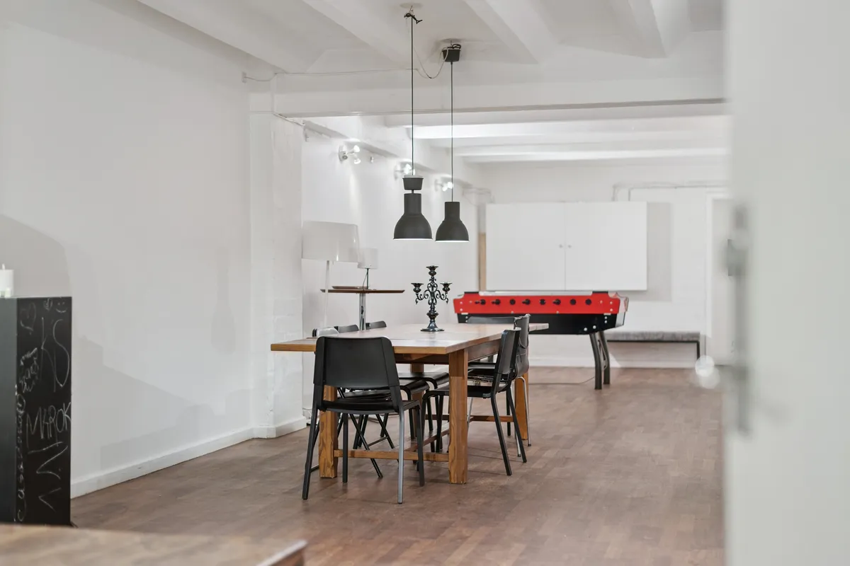 En stor stue med højt til loftet, hvor et langt bord og stolpelampe er i centrum, og en bordfodbold er i baggrunden, alt i et minimalistisk og hvidt farveskema