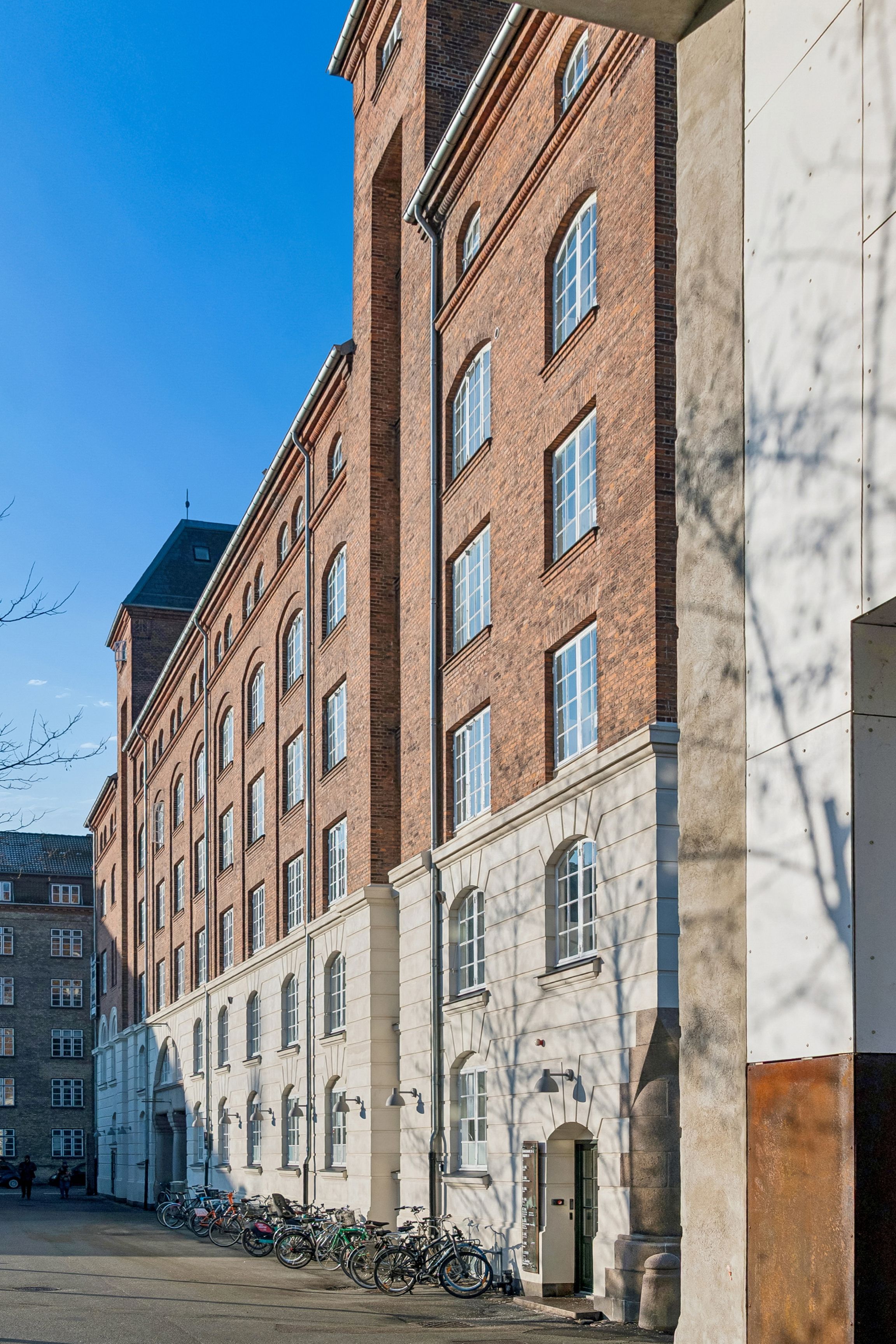 Lyse kontorlejemål i råt erhvervsmiljø med store vinduer og røde mursten. Bicycler parkeret i rækker foran bygningen. Høj gule væg og lysegrå gulv.