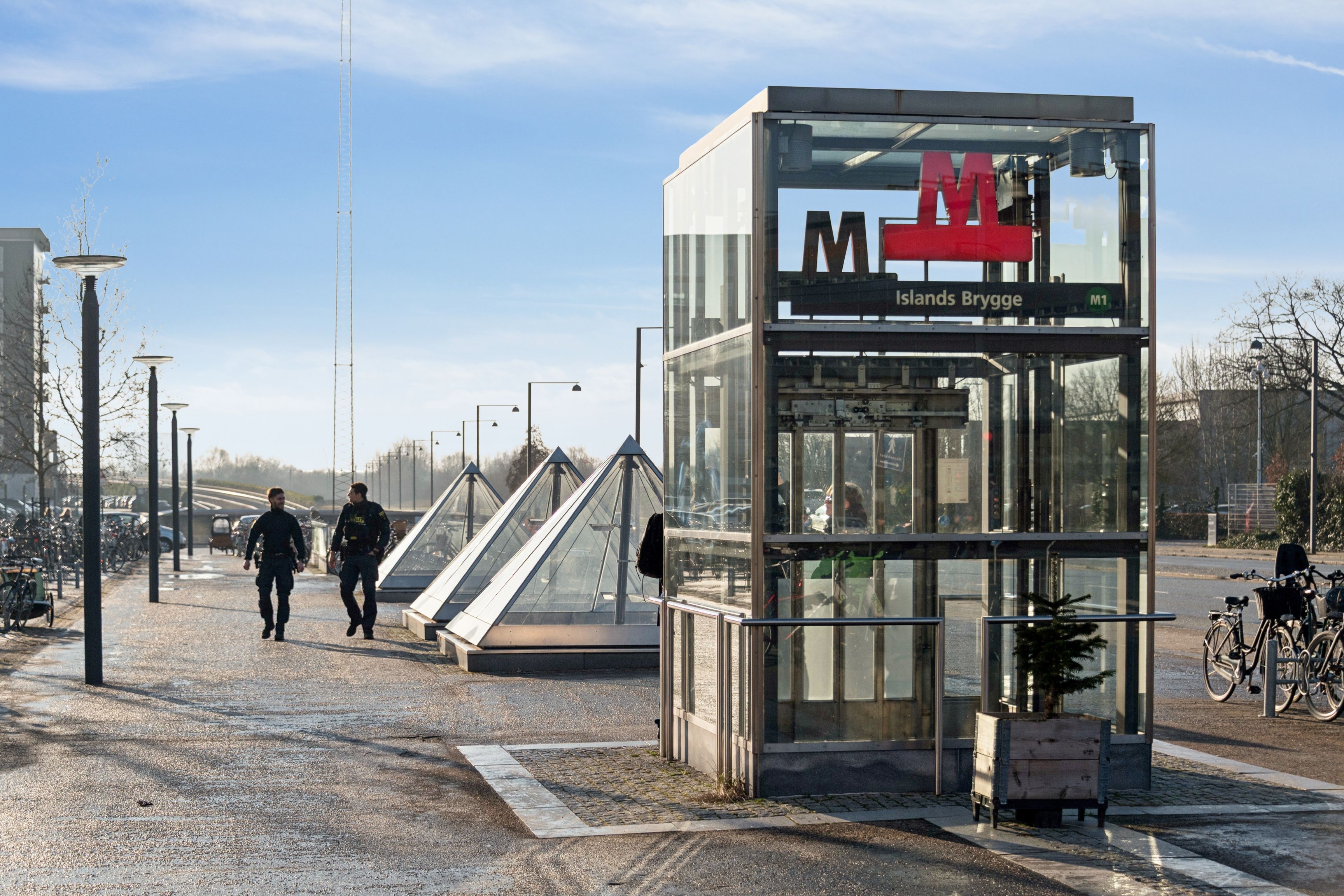 To mænd går gennem et busstoppested ved Islands Brygge med glassdør og M-skilt. Der er flere pyramider og søjler langs vejen.
