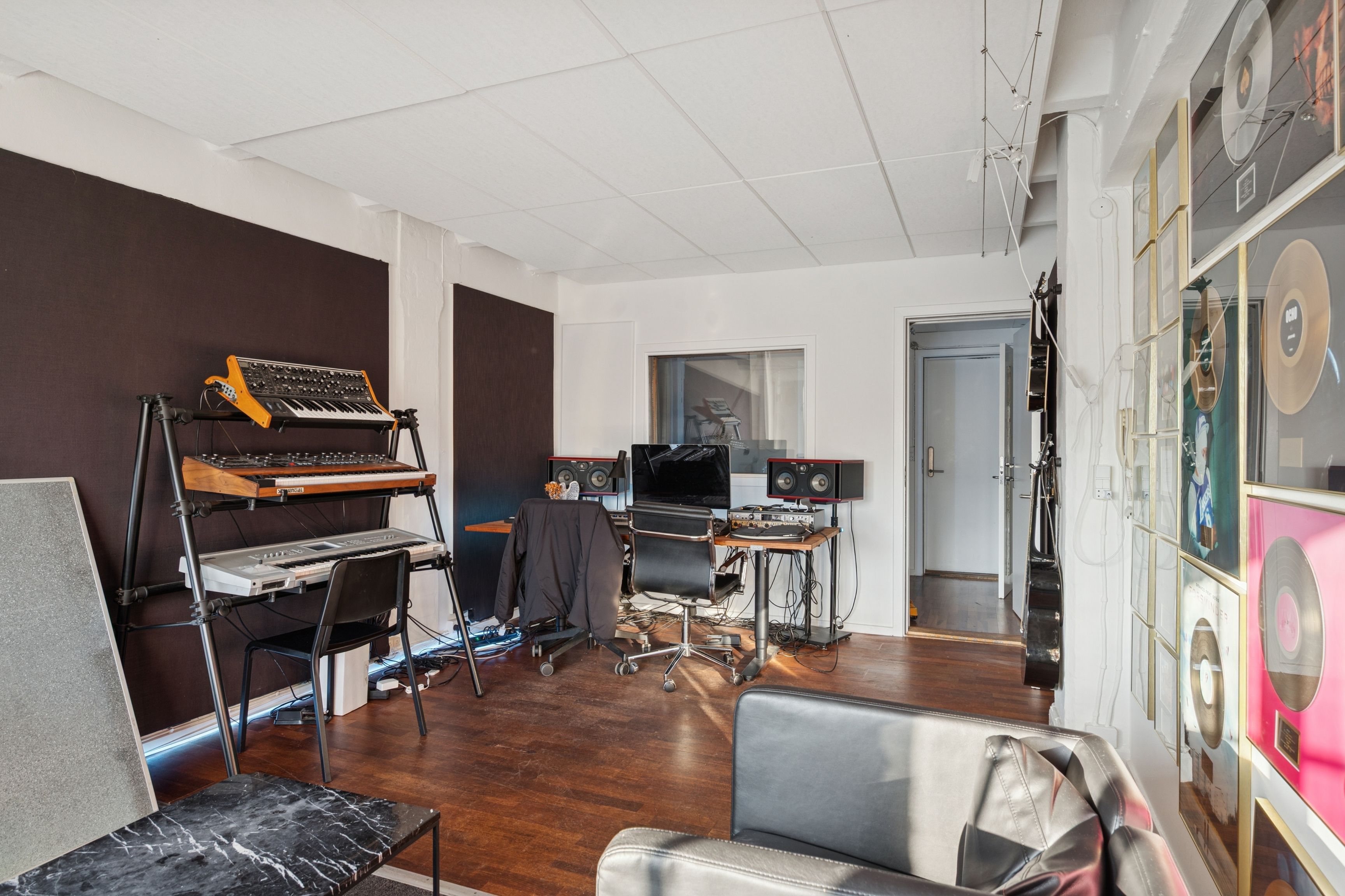 Musikrum med to synthesizere og klavere samt to stolpemonterede højtalere og en bærbar computer. Det er en lukket dør i baggrunden og flere vinylplader samt musikinstrumenter på væggen