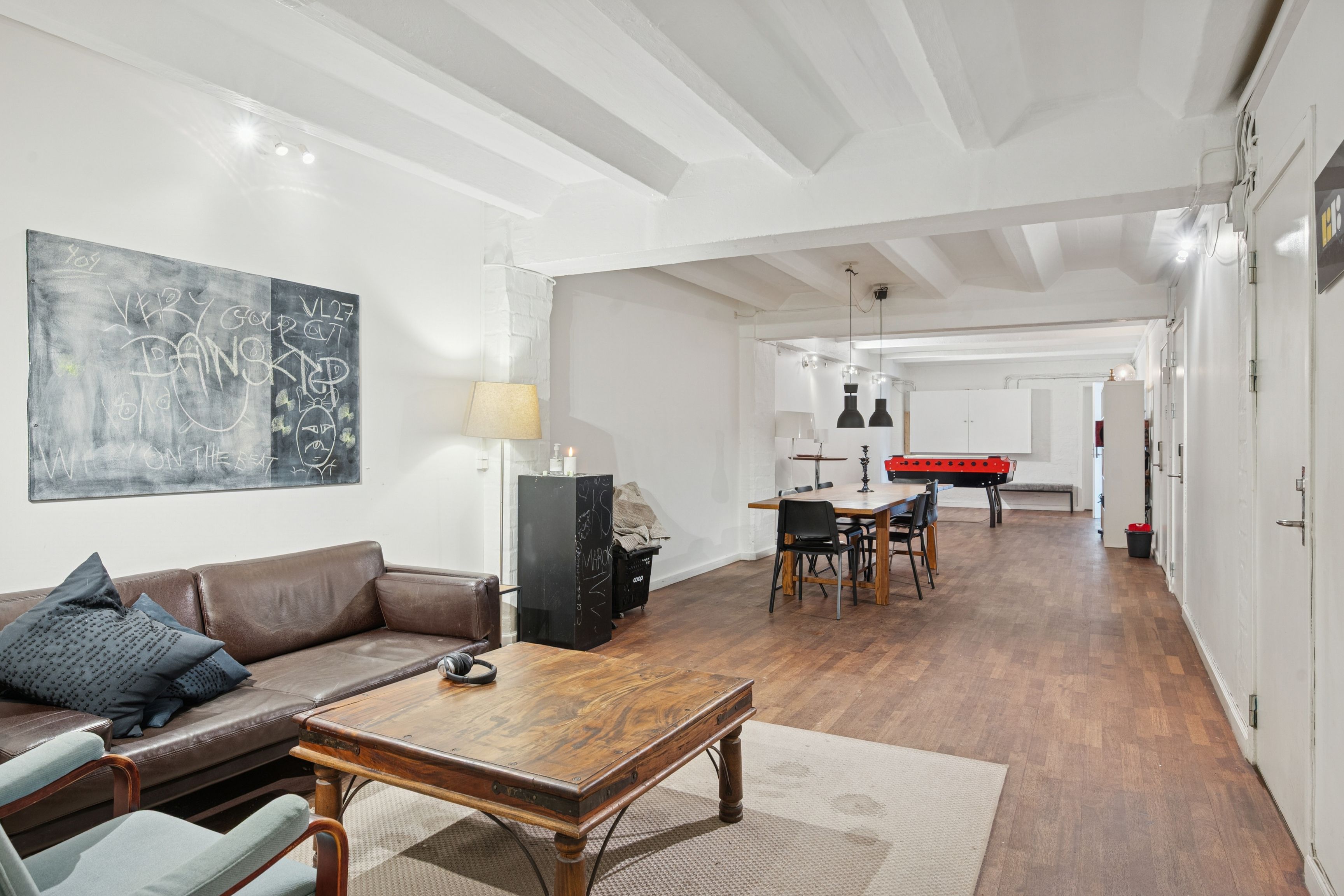 Kontorrum med loftsbjælke, sofa, skrivebord og spisestue med poolbord og lysekroner. Væg med skrift og lysepunkter og loftsbjælke med skråtage