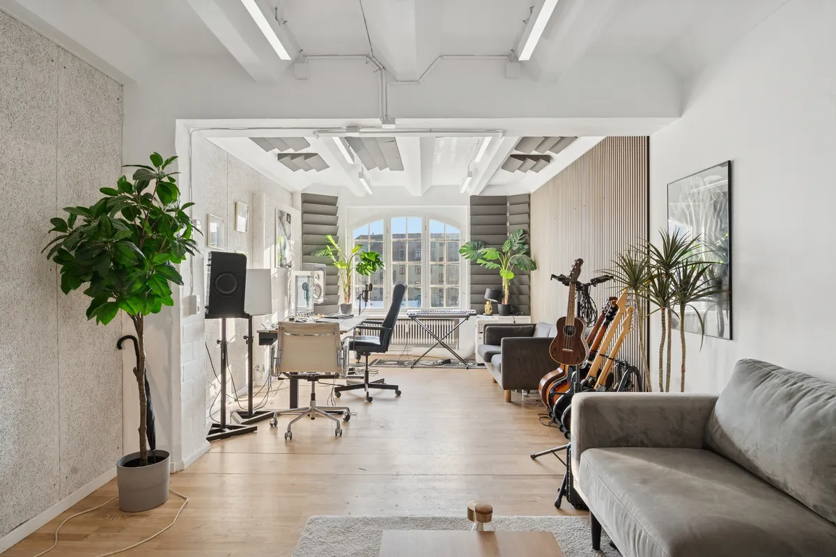 Kontorrum med højt til loftet og hvide vægge, hvor musikinstrumenter står på gulvet og en stor sofa. Der er en lysekron og plante i hjørnet.