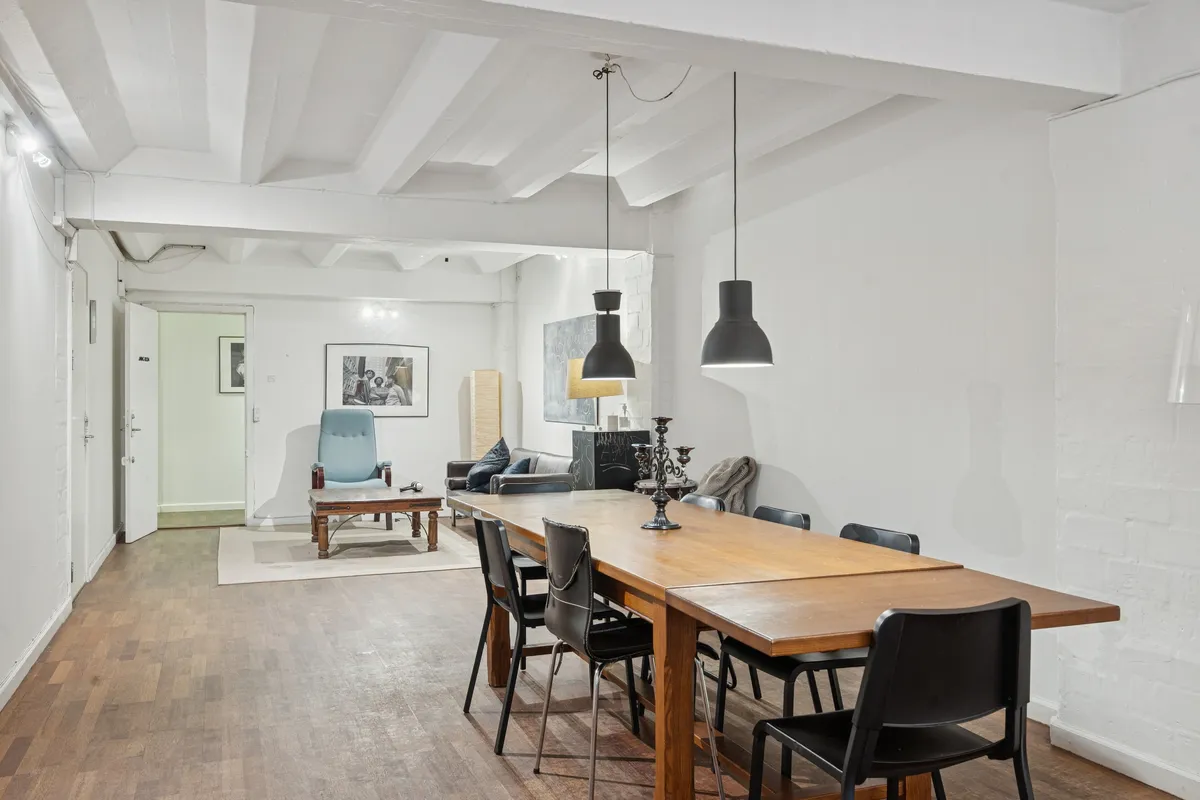 Lyst og industrielt kontor med massiv træbord, sort og blå stolpe, loftsbjælker, lysekroner, fotorammer og en hyggeligt indrettet stue med sofa og lampe.