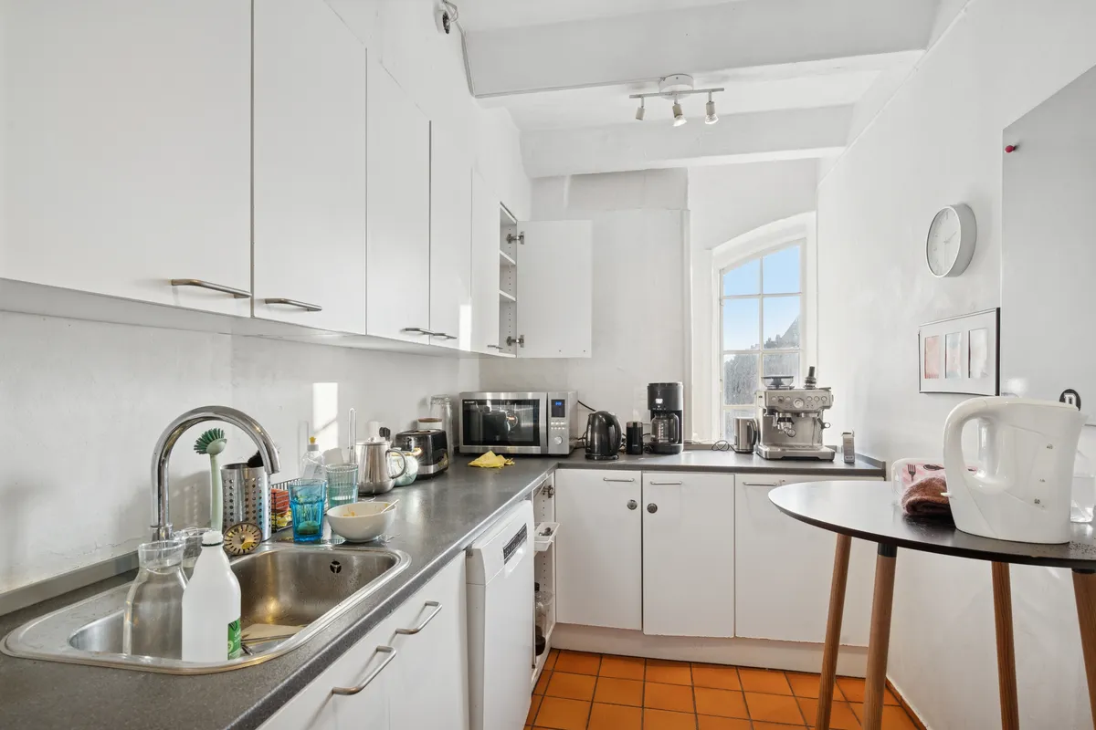 Lys moderne køkken med hvide skabe, lysekroner, tørretumbler, kedel, mikroovn, kaffemaskine, køleskab og vaskemaskine samt vinduesset og åbne skabe.