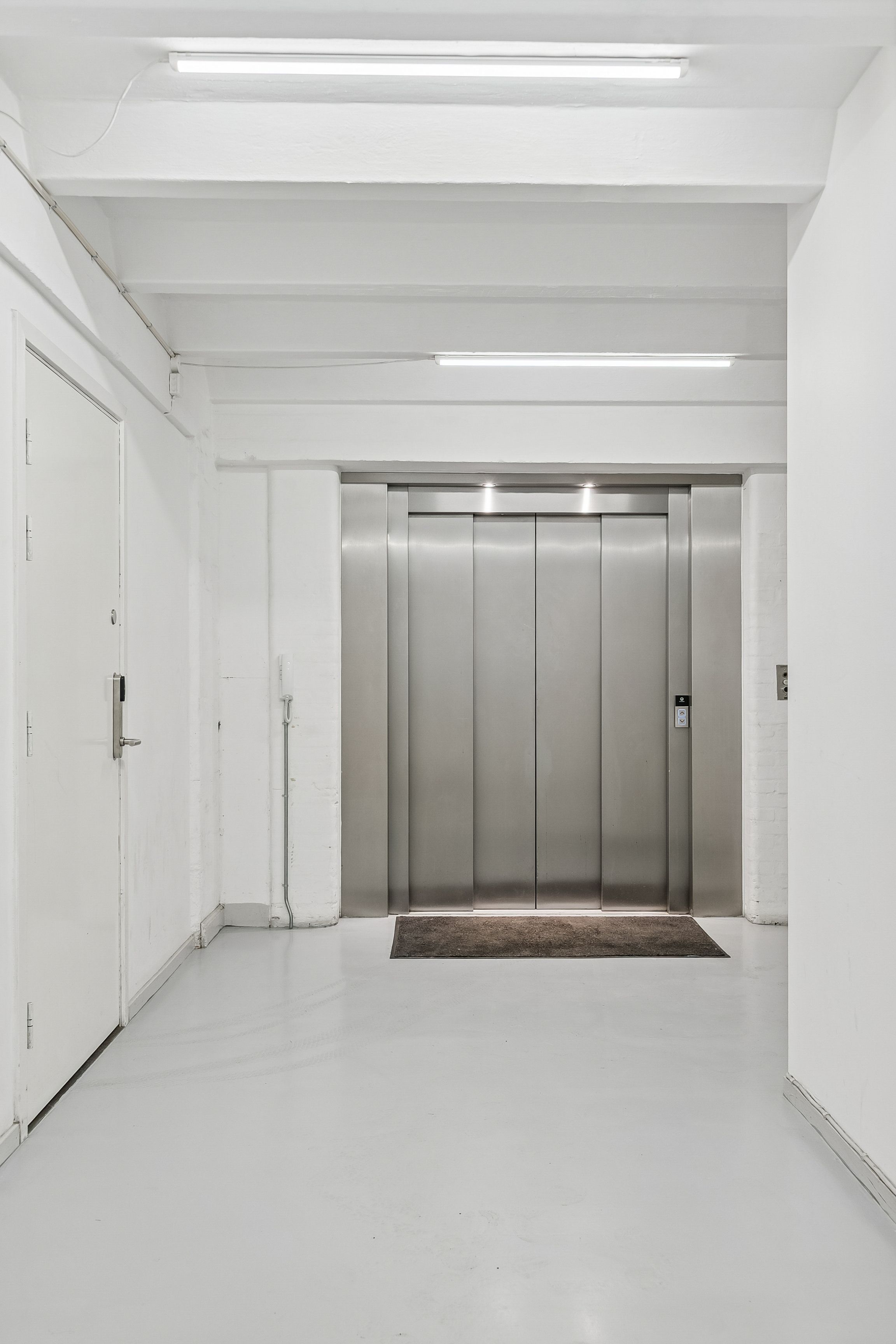 Rummelige lokaler med hvide vægge, moderne gulv og to elevatorer i et industrielt miljø med en brun tæppe foran elevatorerne og hvide loftsbelægninger med lyse striper