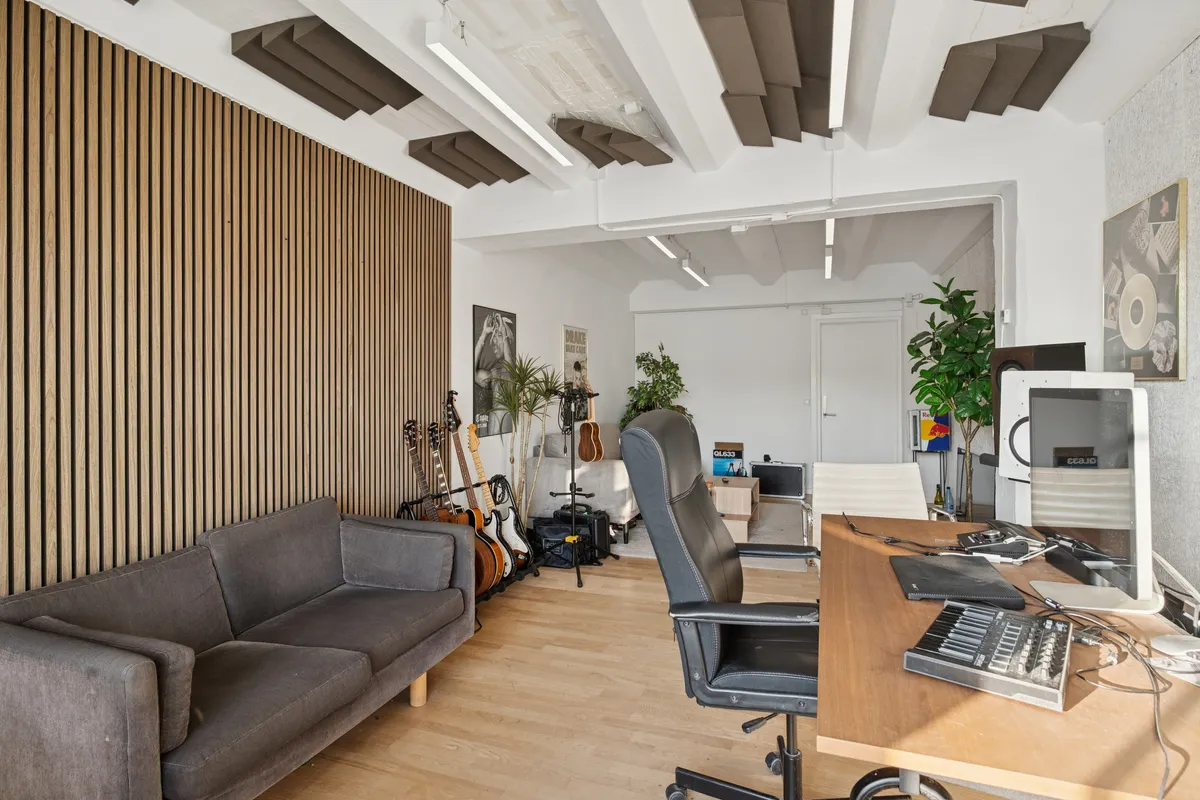 Kontorrum med lyse hvide vægge og træpaneler, møbler og musikinstrumenter, stol, skrivebord, computer, plante, monitor og lydsystem