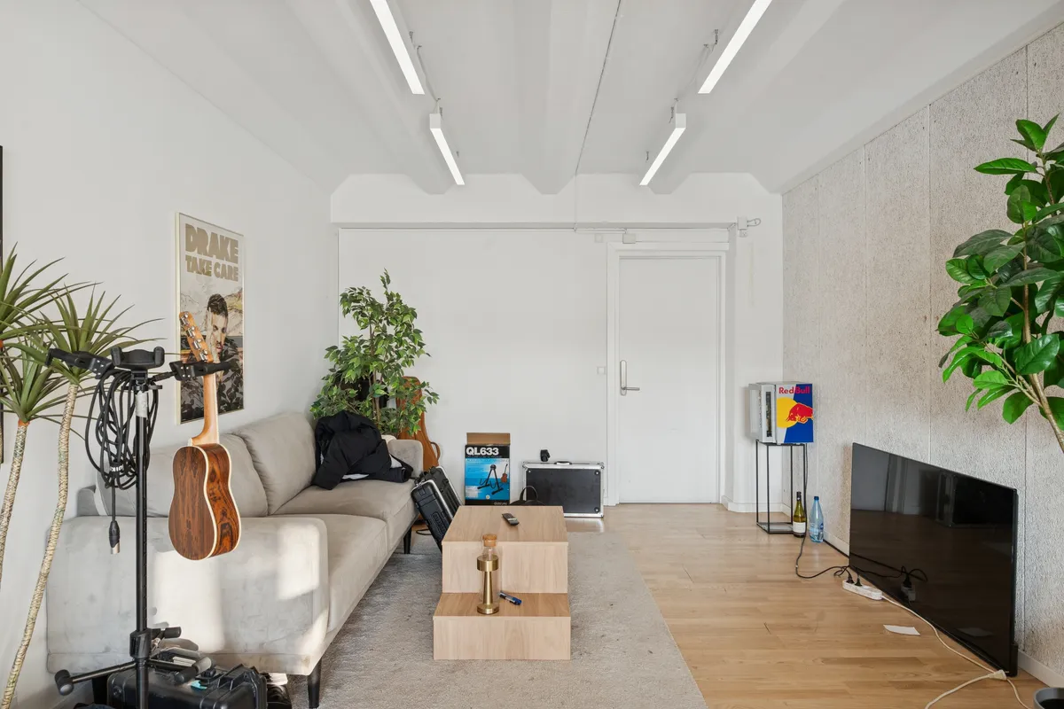 Kontorlejemål med højloft, moderne møbler og hvide vægge, samt en stor sofa, træbord, tv og flere plantager