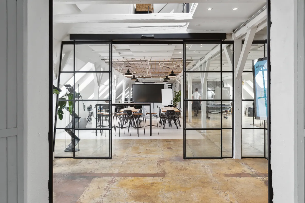 Kontorrum med åbne glasdøre og højloft. Rundt i rummet står lyse, moderne stole og borde. Der er et projektionsapparat og en person i hvid skjorte i baggrunden.