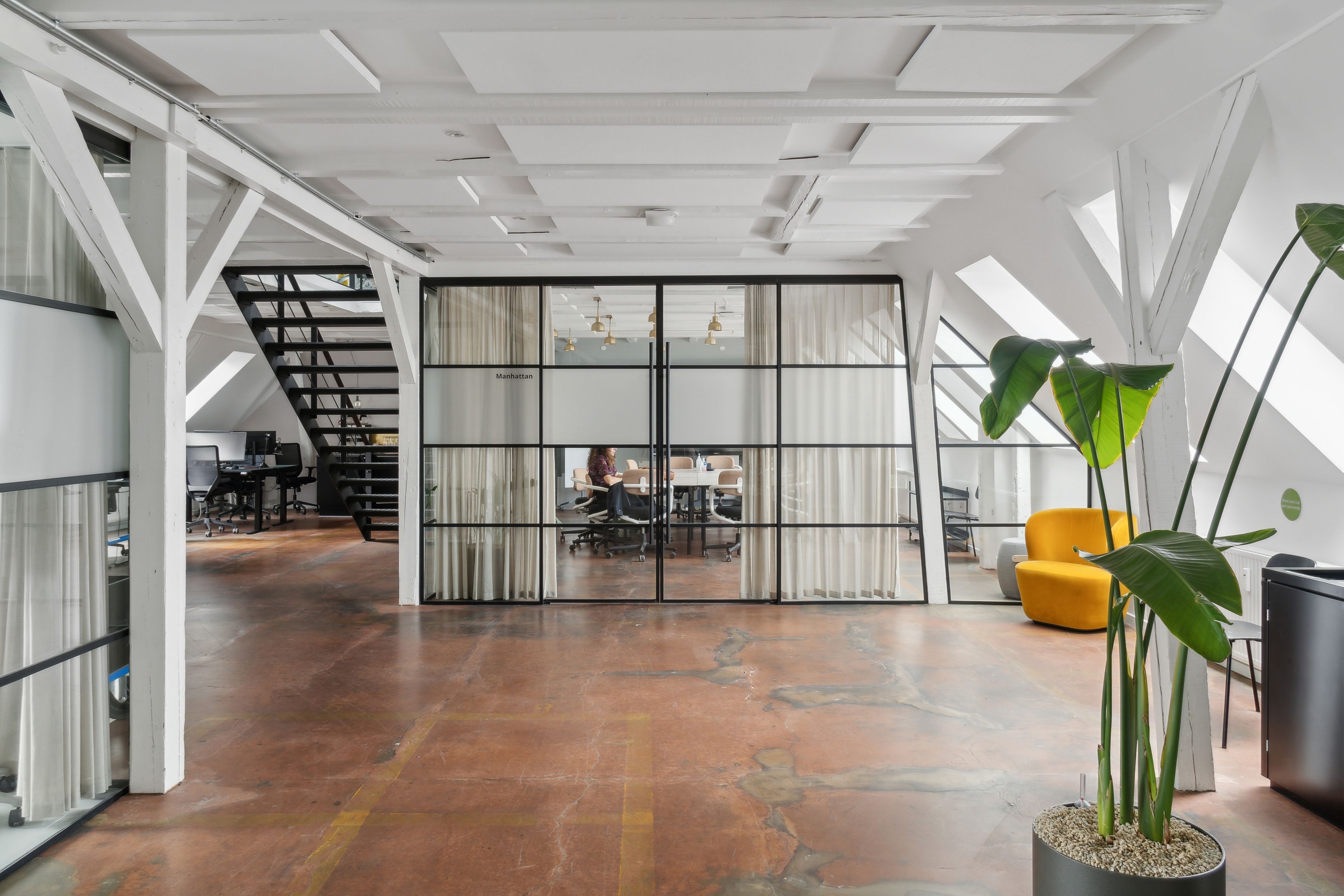 Rå og charmerende kontor med loftsrum, møbler og plante på Manhattan. En person sidder ved et bord. Loftet er hvidt med bjælker.