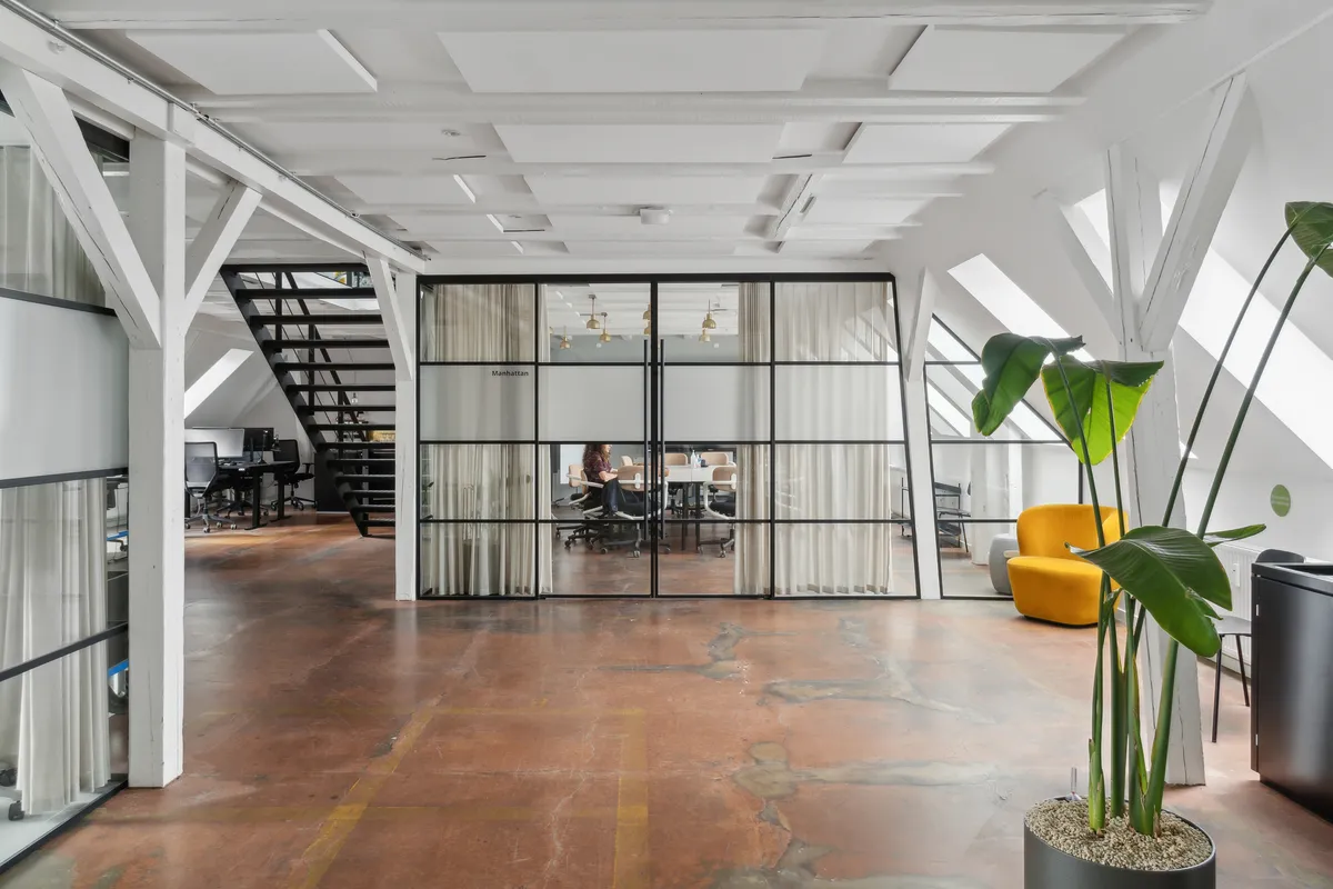 Rå og charmerende kontor med loftsrum, møbler og plante på Manhattan. En person sidder ved et bord. Loftet er hvidt med bjælker.