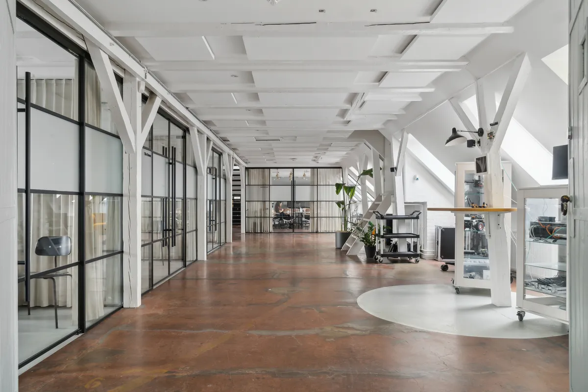 Kontorområde med rå lofter, glasvinduer og lysegulve. Møbler og plante i rummet. Et stort skrivebord med computerudstyr. En stol og et bord i enden af korridoren.