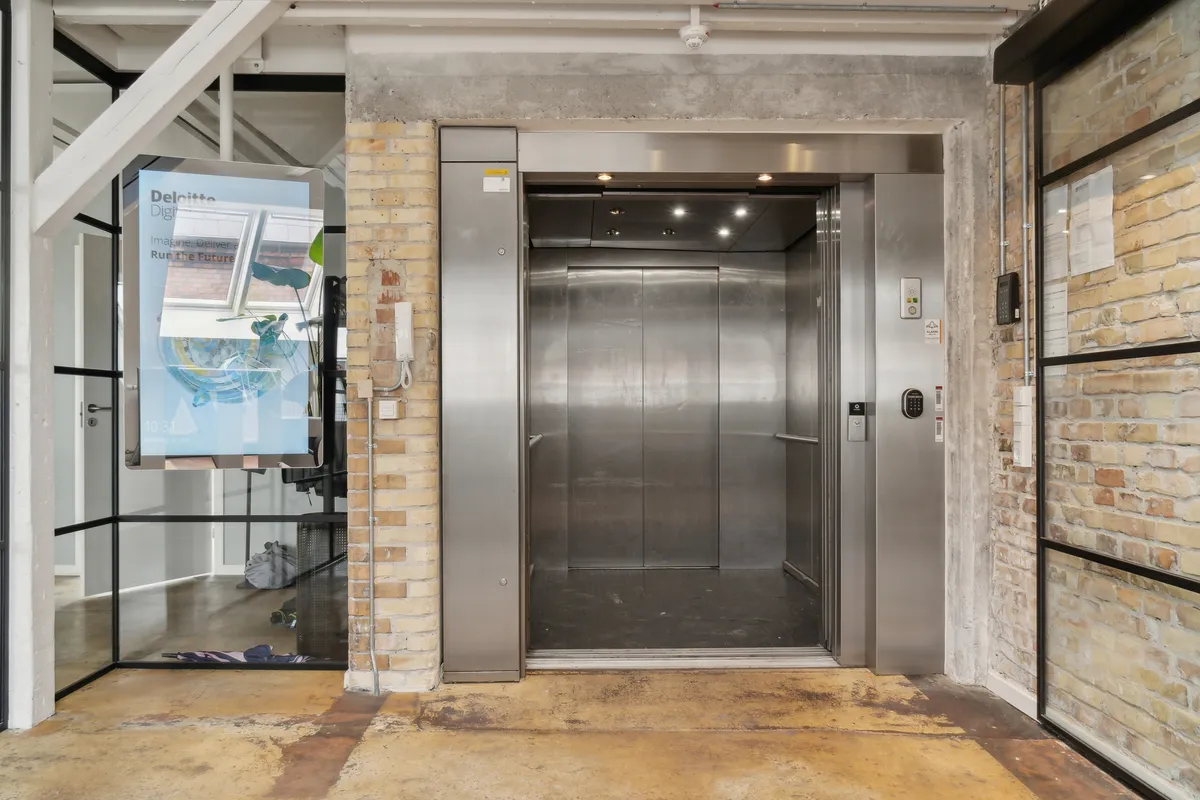 Indkørsel til et kontor med åben elevator og glasdør mod en mur af røde mursten. Der er en digital annonce med teksten Deloitte Digital, og der er en røgalarm på loftet.
