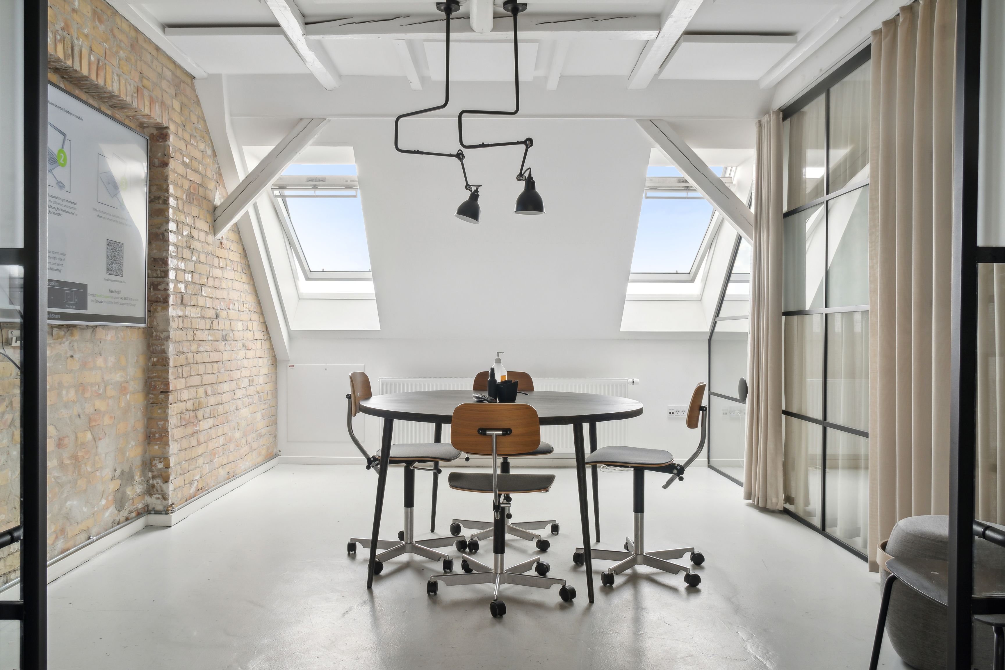 Kontor med rundt bord og fire stolpestole samt loftsvinduer og rørlampe på loftet