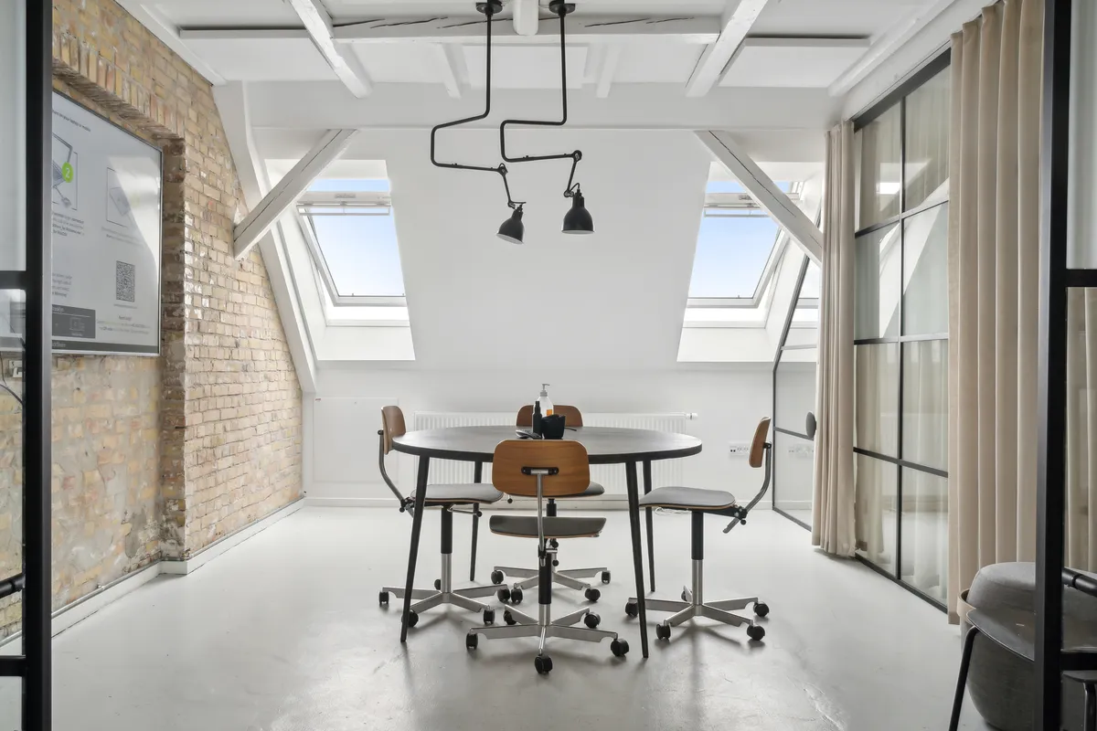 Kontor med rundt bord og fire stolpestole samt loftsvinduer og rørlampe på loftet