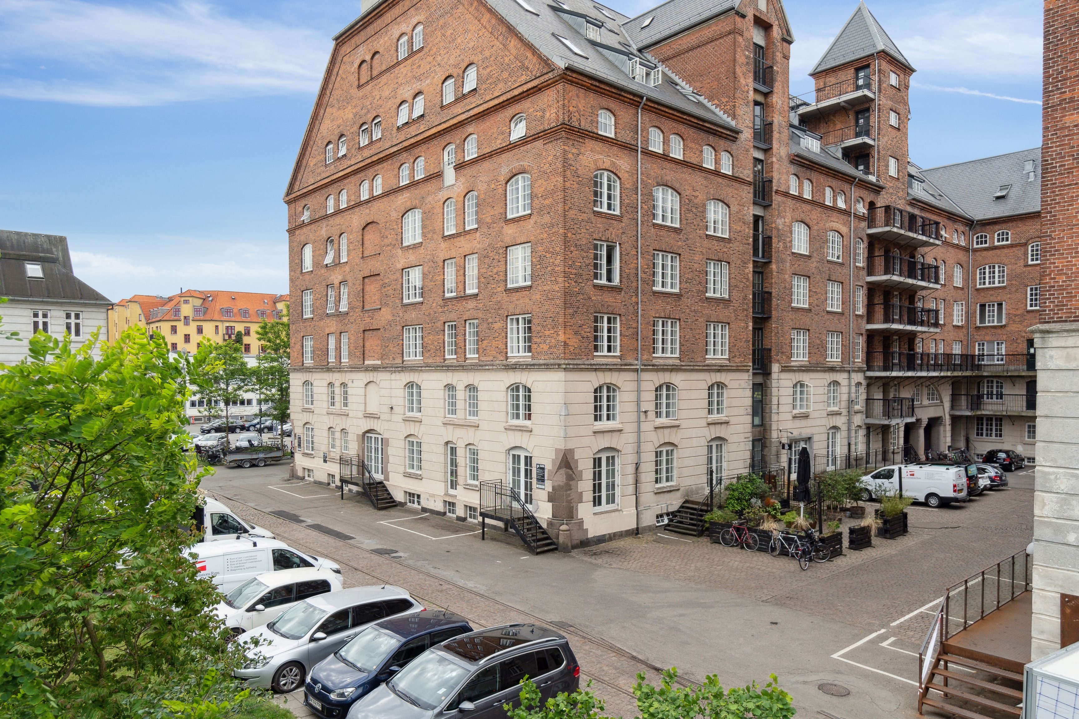 Kontorlejlighed med rå detaljer og kreativ atmosfære til leje i København. Historisk rødt murstenshus med terrasser og parkerede biler. Planter og cykler parkeret udenfor.