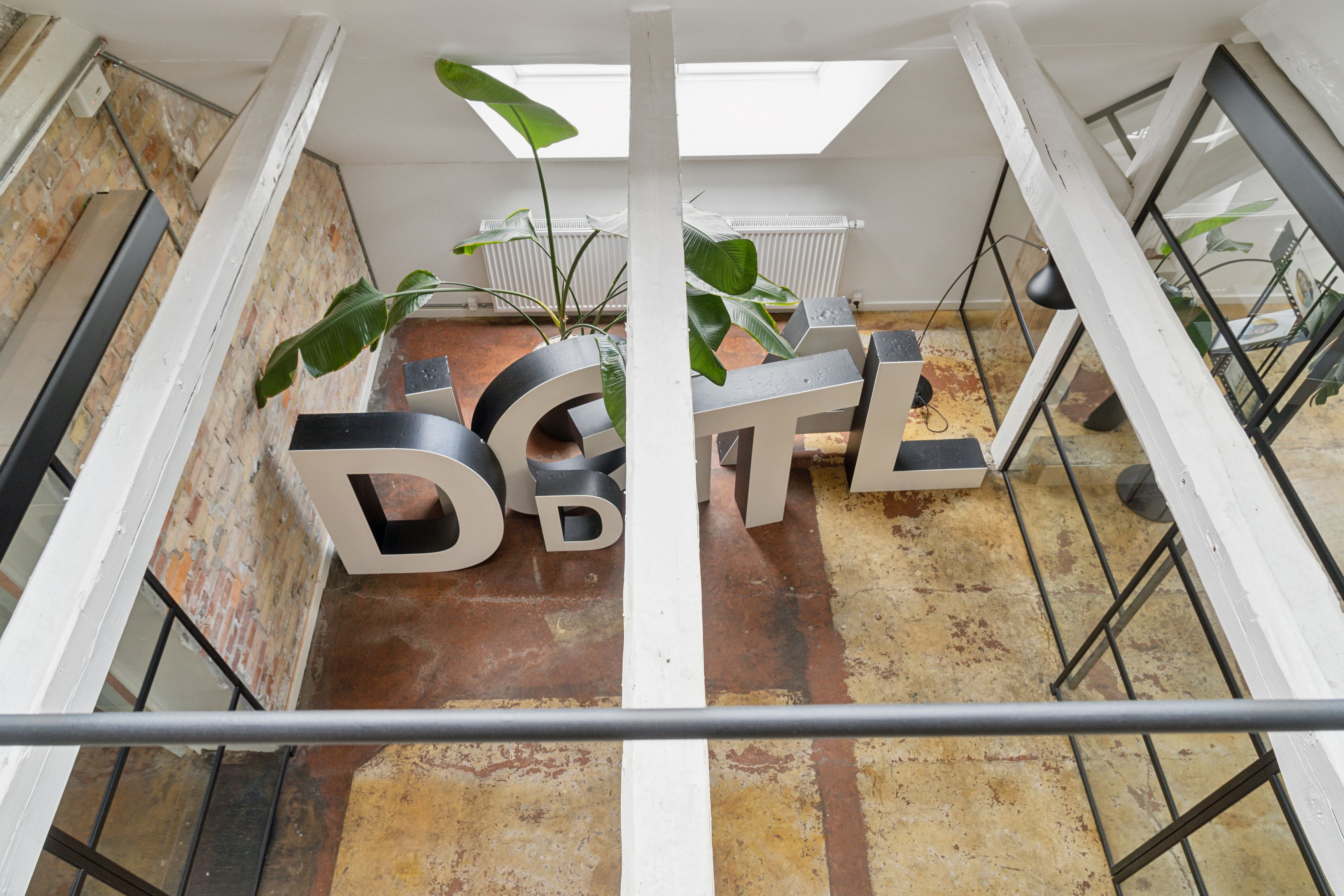 Store bogstaver med DGT-mærke står på gulvet i en loftslejlighed med plante og gulvtæppe samt møbler og vinduer i baggrunden