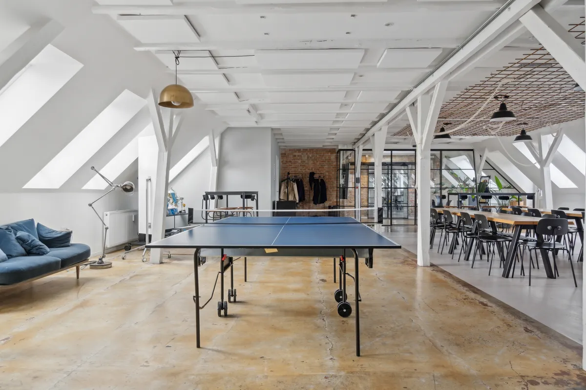 En stor loftsværelse med pingpongbord, lysekroner, hvide bjælker og lyse lofter. Bord med kuglebånd og sofa med puder og en hvid radiator.
