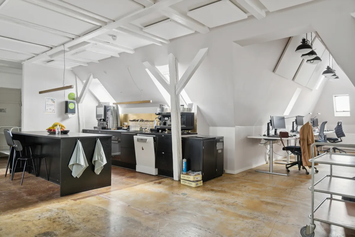 Kontorloft med loftsvinduer og loftsbjælker, lysekroner og lysepunkter, køkken med kaffemaskine og udstyr, skriveborde med computere og skriveborde, trægulve, to stole og hængekøkkenhåndklæder.
