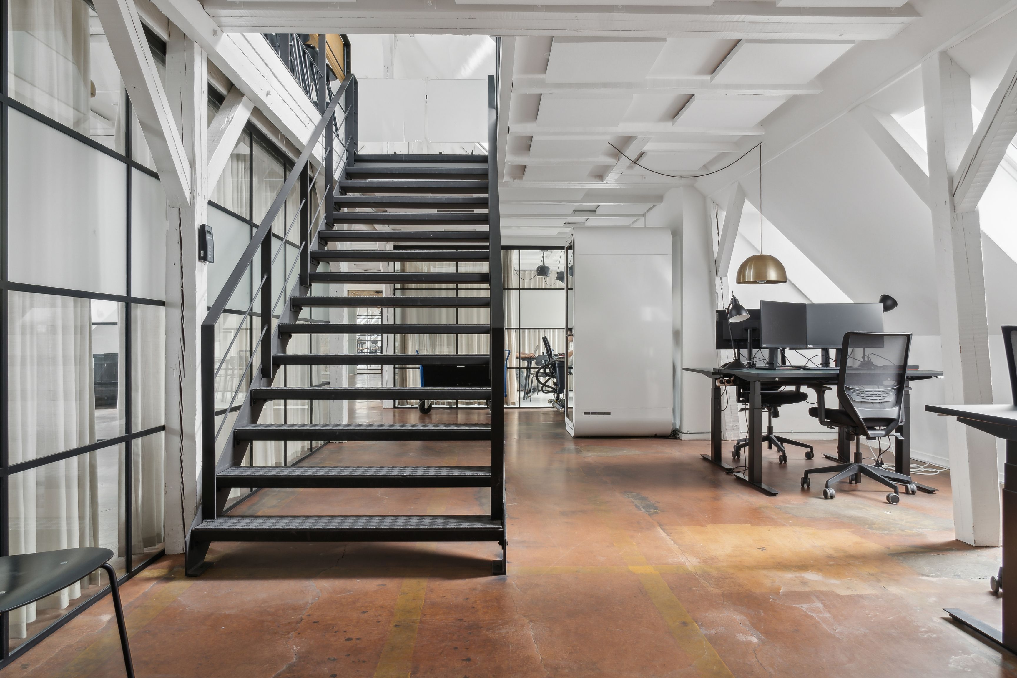Kontorområde med rå loftsstrukturer, høje vinduer og minimalistisk møbelsætning med skriveborde og stole