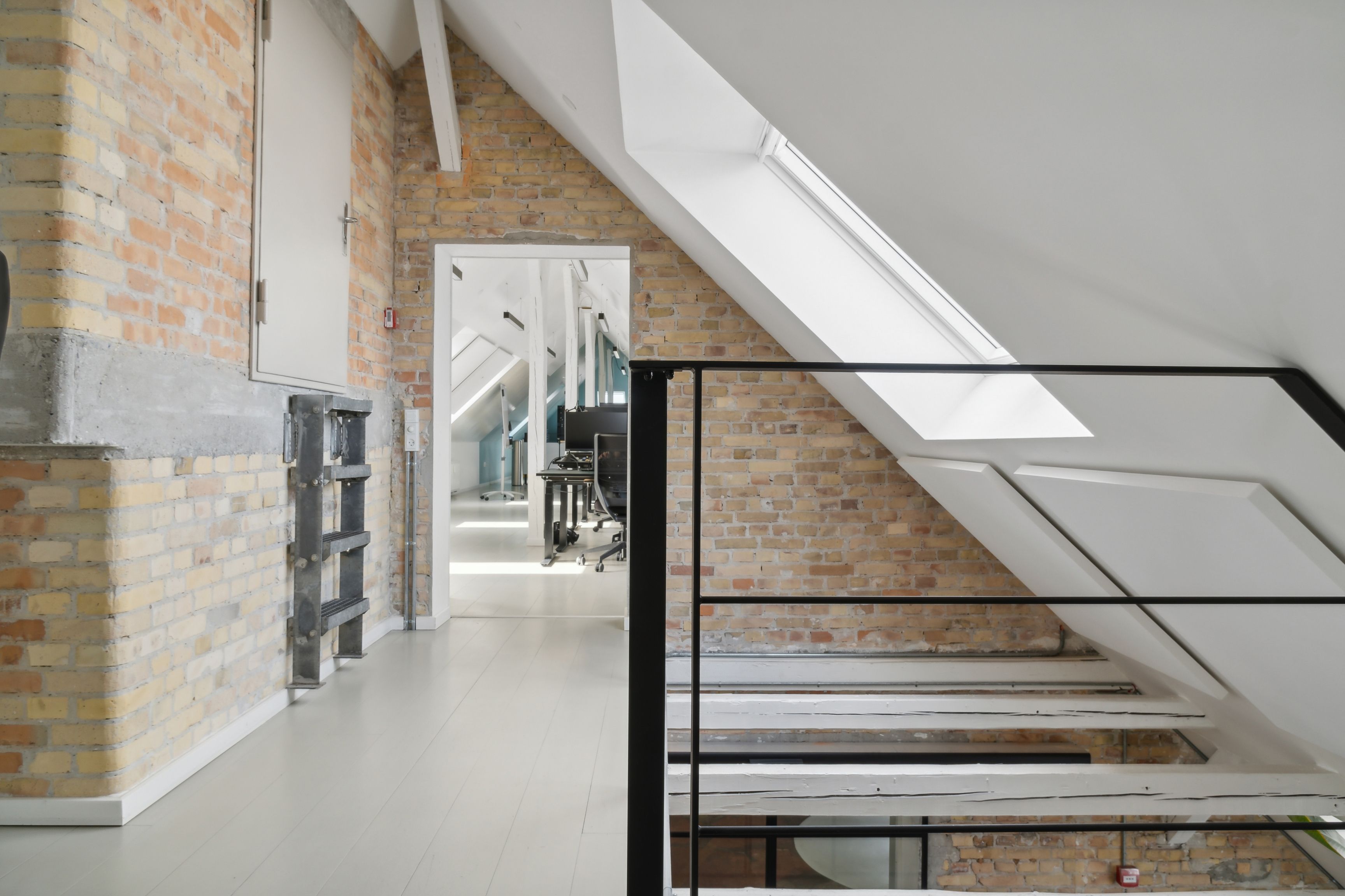 Kontorlokaler med loft og rå røde mursten. Stiger med metalgevær og hvidt loft. Kontorbord og computer er i rummet.
