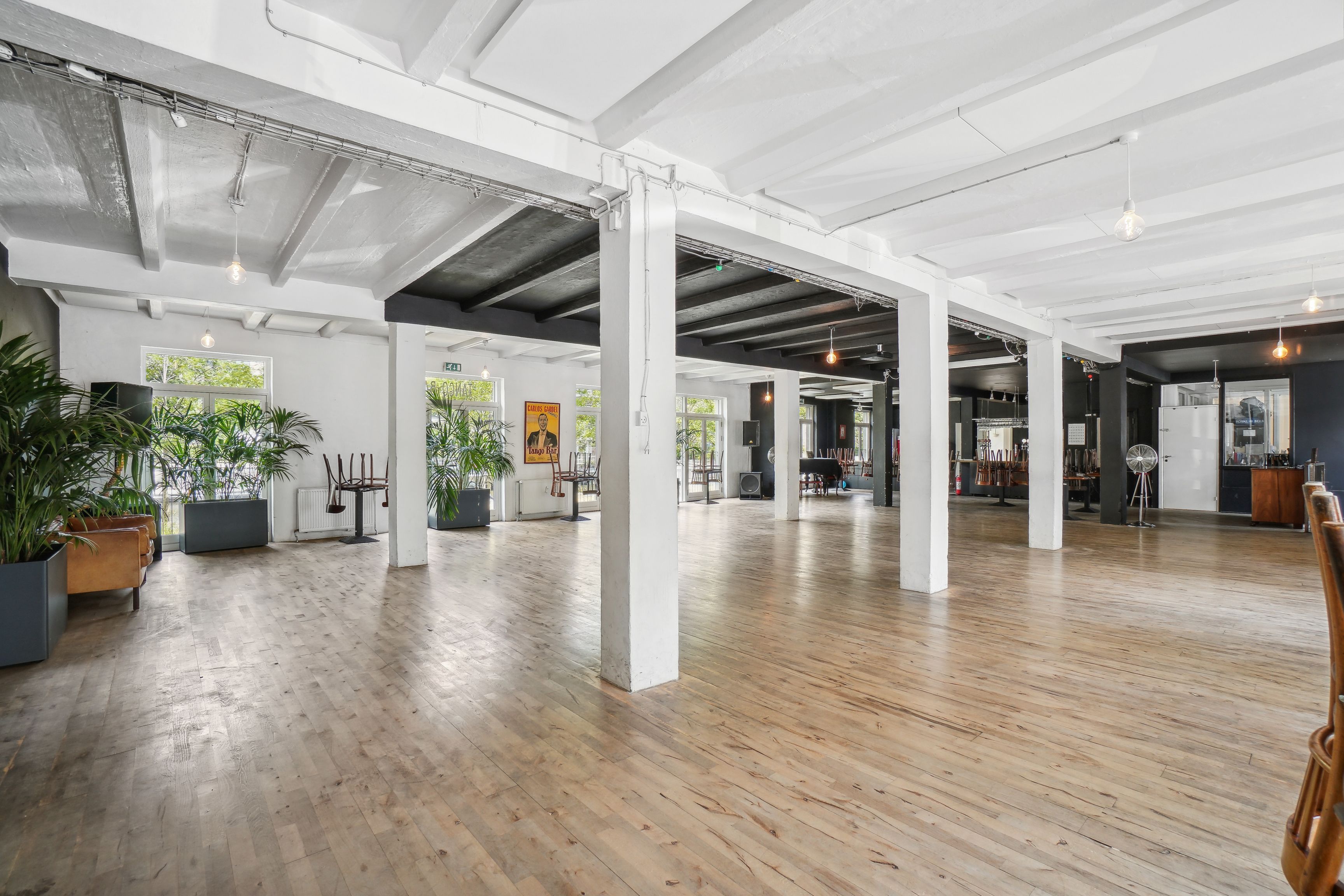 Rå og charmerende erhvervsmiljø i Njalsgade-kvarter. Store, lyse rum med trægulve, loftsbjælker og hvide vægge. Planter og møbler er placeret rundt i rummet. To store vinduer med udsigt til grønne træer.