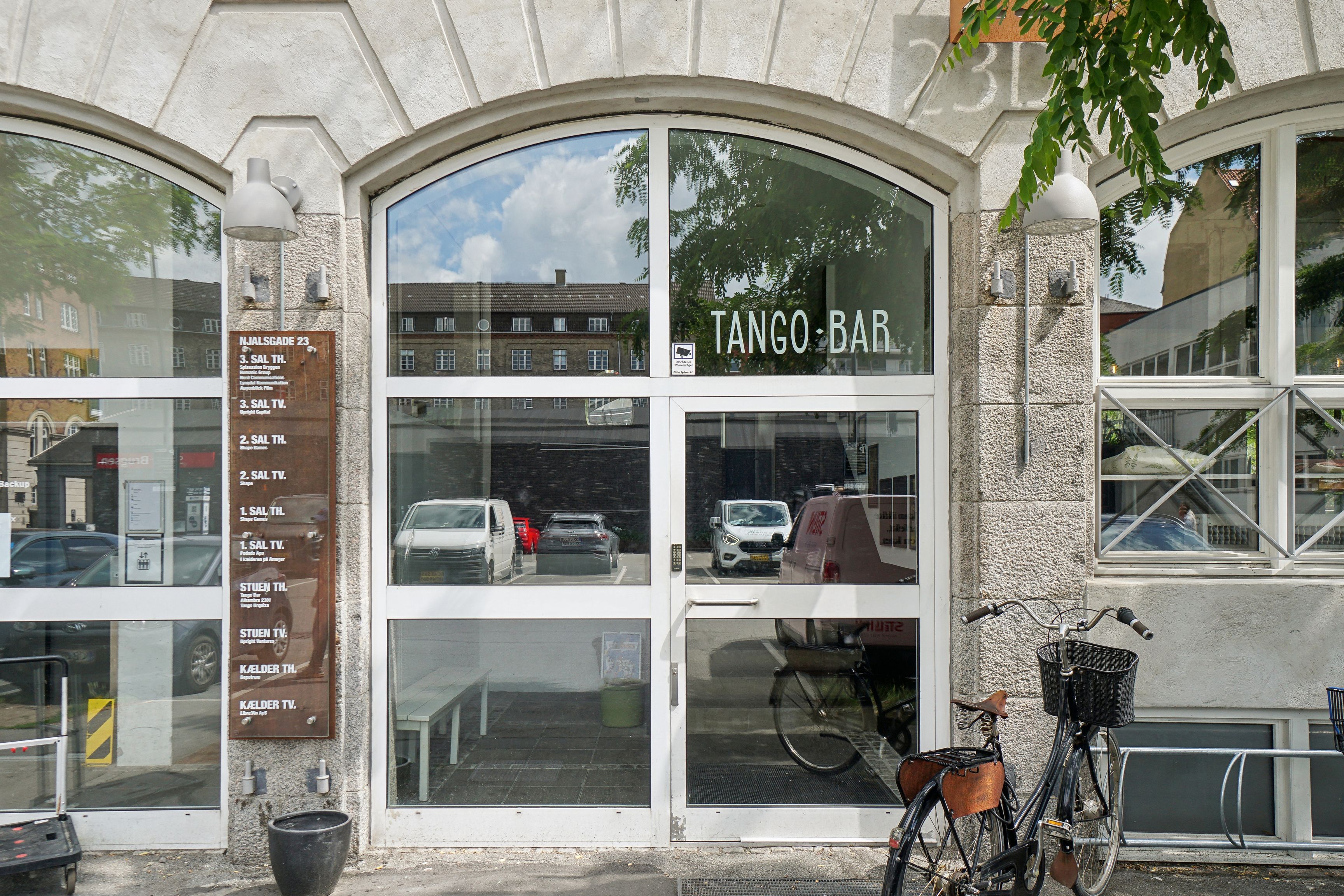 Bygning med glasruder og hvide vægge og teksten Tango-bar på glasdøren og en cykel parkeret på forsiden med en kurv og en kasse på cykelsædet