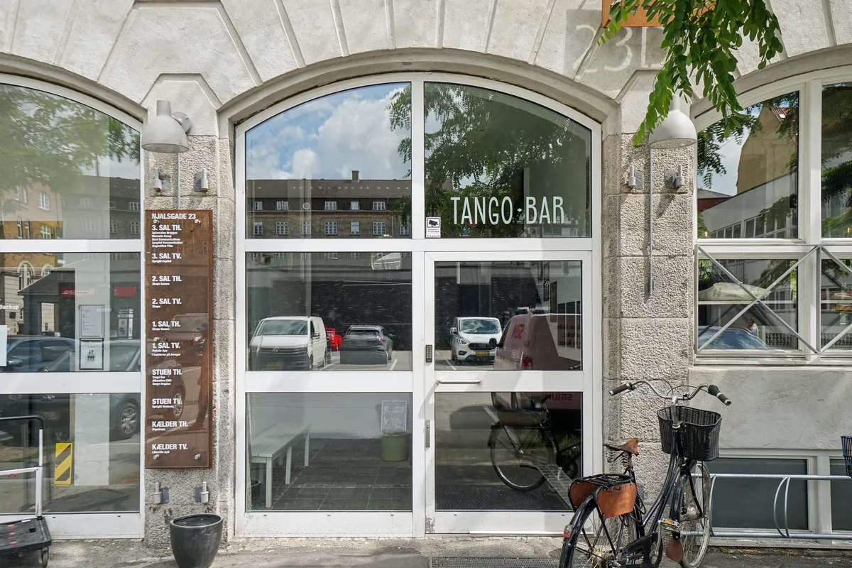 Bygning med glasruder og hvide vægge og teksten Tango-bar på glasdøren og en cykel parkeret på forsiden med en kurv og en kasse på cykelsædet