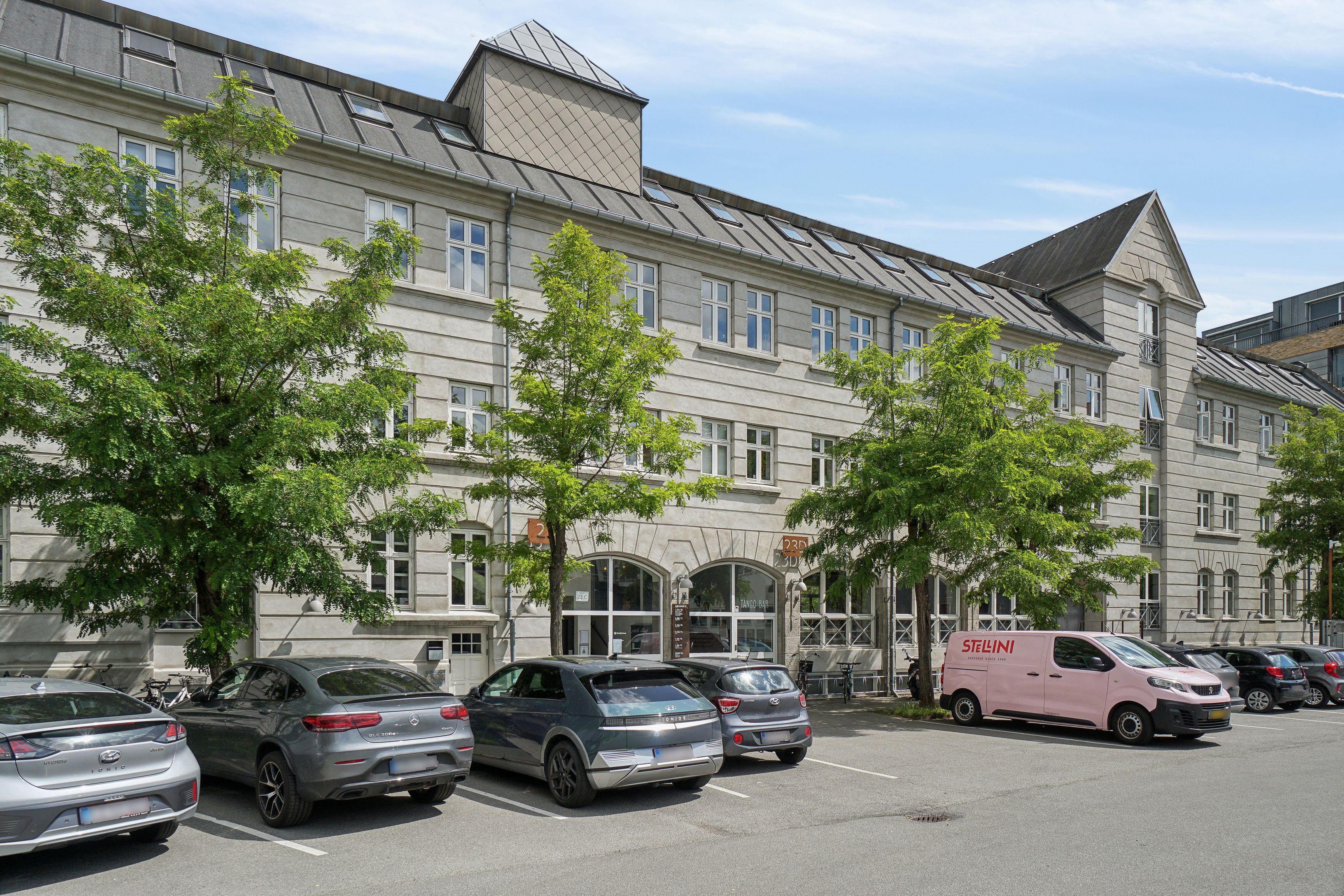 Steder med flere biler parkeret foran Njalsgade 23C. Bygningen har flere vinduer og et pinkt Stelini-leveringsvogn. Foran er flere træer, og en stel er parkeret ved siden af bygningen.