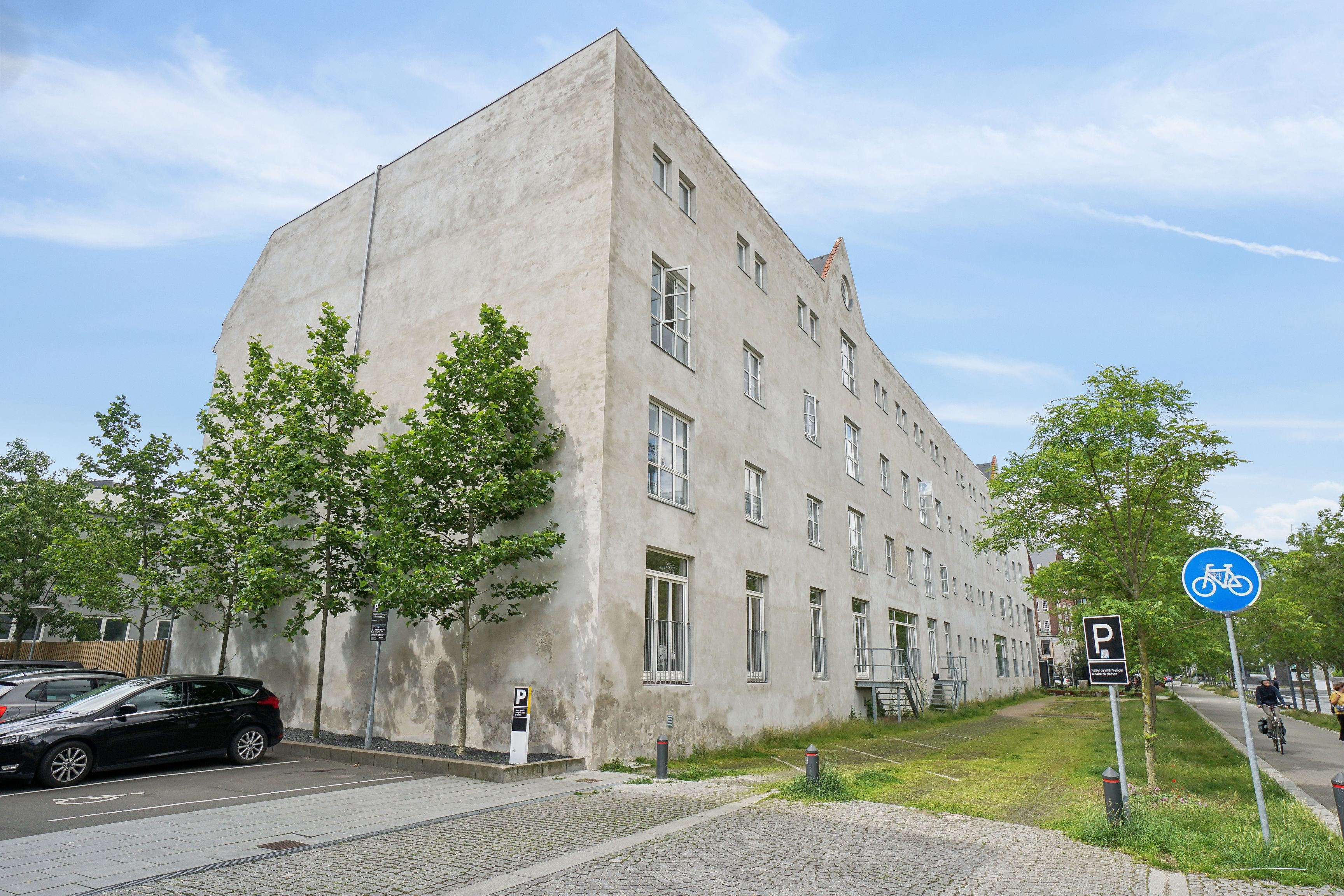 Et lysegråt hus med flere vinduer og parkeringspladser foran i Njalsgade 23C, 2300 København S. En person cykler forbi på cykelstien med skovløb og træer.