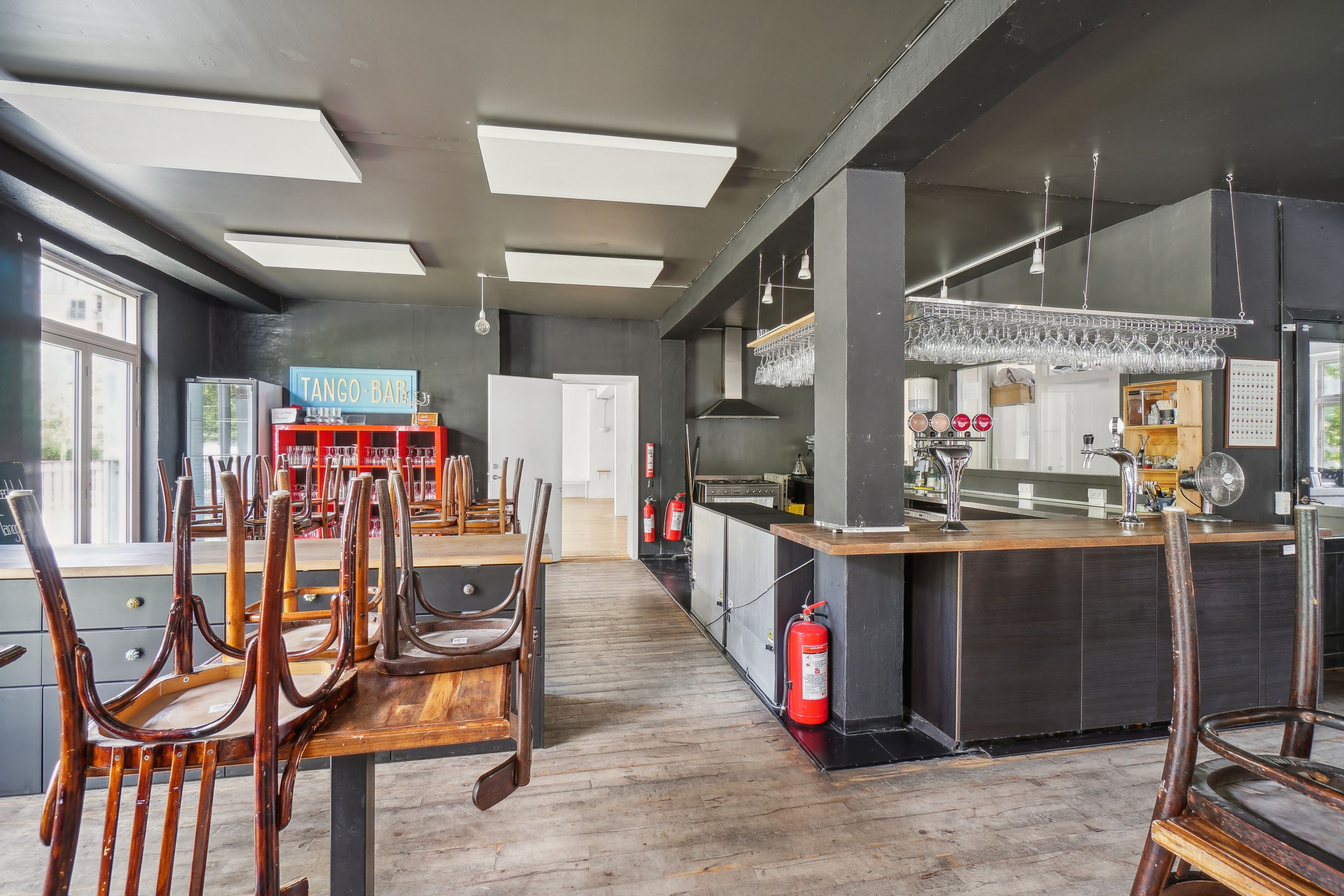 Inde i et værkstedslokale med trægulve, flere stole og borde, vinglasreol og en stang med vinglas, et rødt reol med glas og et køleskab og en rødt brandvandsslukker.