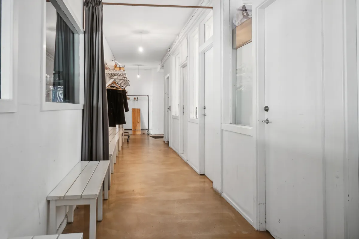 Langt, smal hall med hvide vægge og lysegul gulvbelægning. To hvide bænke står mod højre væg og tøjstativ står mod venstre. Bag tøjstativet er der en klædeskab.